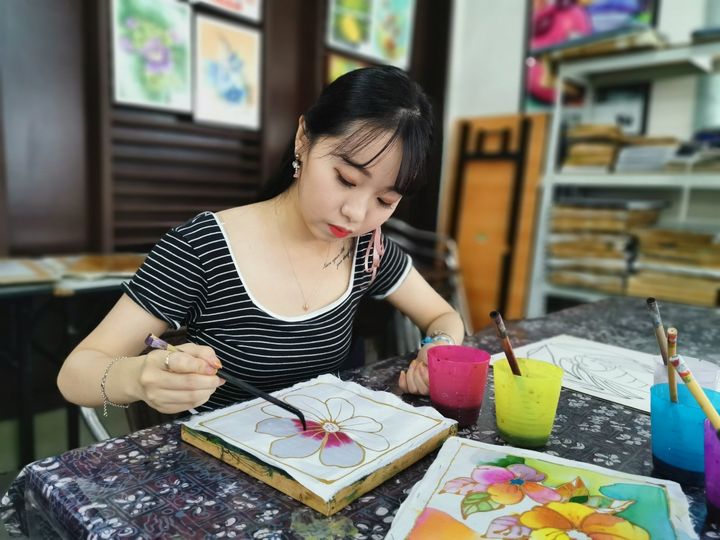 Woman painting a floral design with a paintbrush, sitting at a table with art supplies, indoors.
