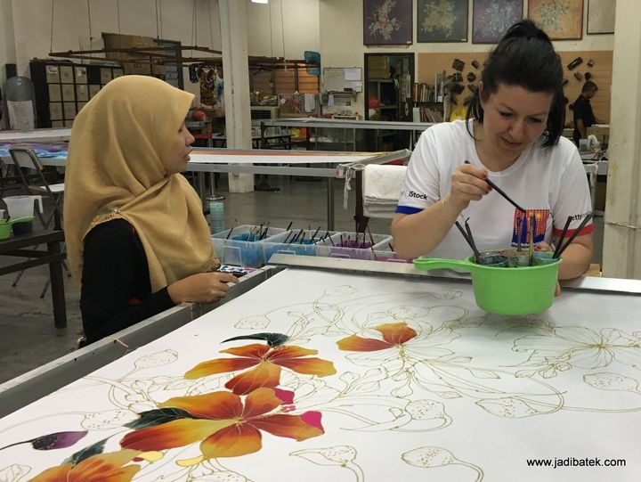 Two women painting batik artwork on a large table in a workshop. One wears a hijab.