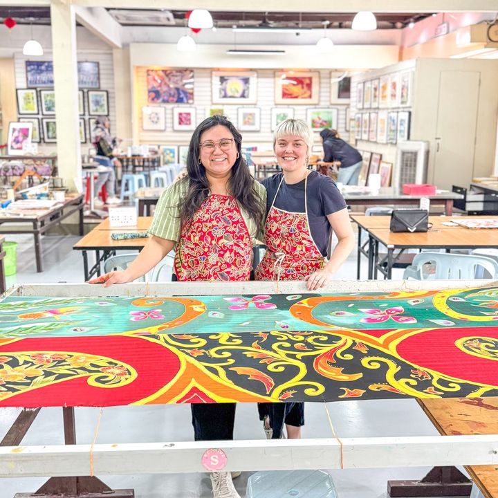Two women in aprons pose with a vibrant batik painting in an art studio.