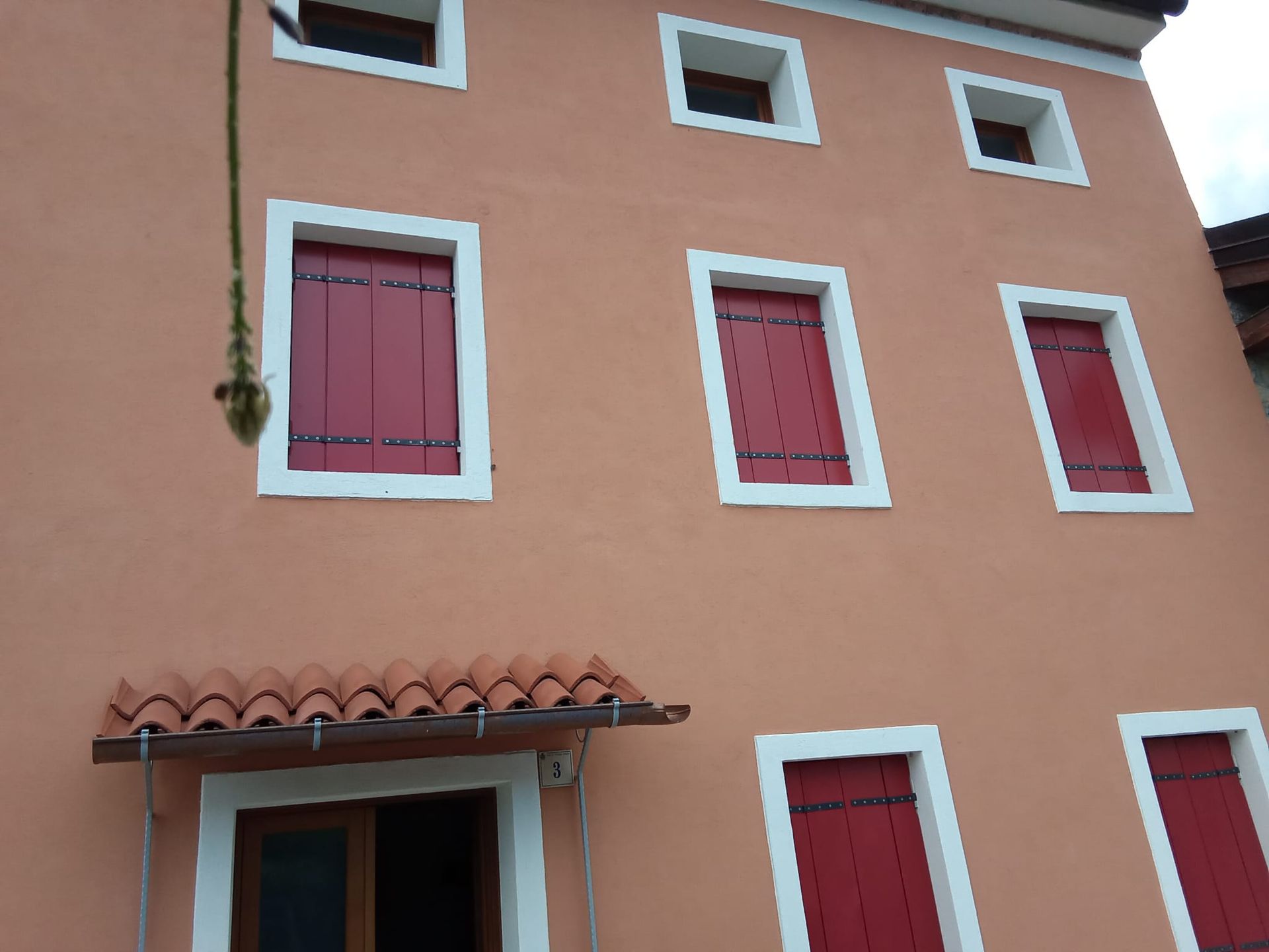 Edificio in stucco rosa con finestre con persiane rosse e una tettoia di tegole sopra la porta.