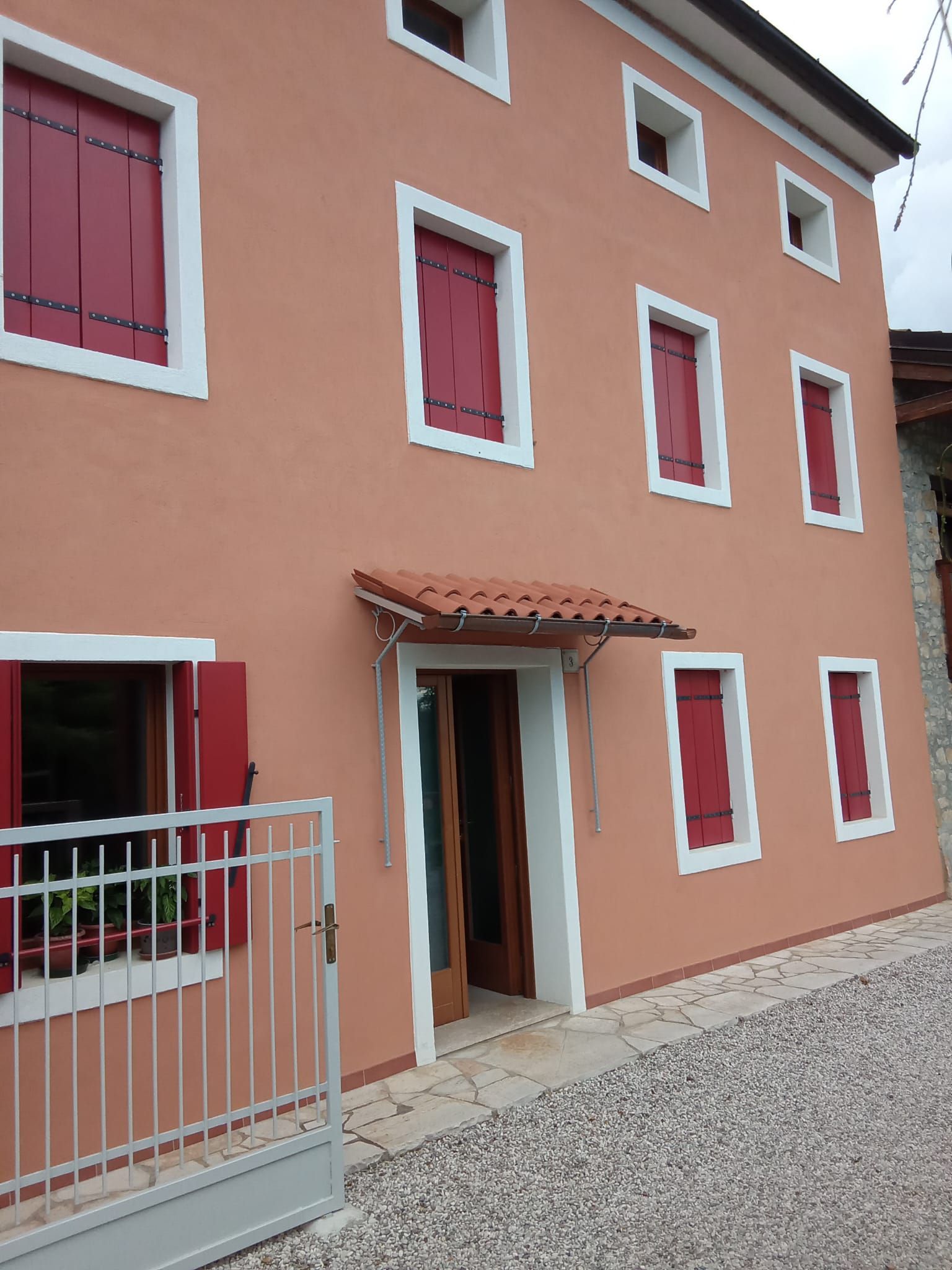 Facciata della casa color pesca con finestre incorniciate di rosso, una piccola tenda da sole sopra la porta e un vialetto di ghiaia.