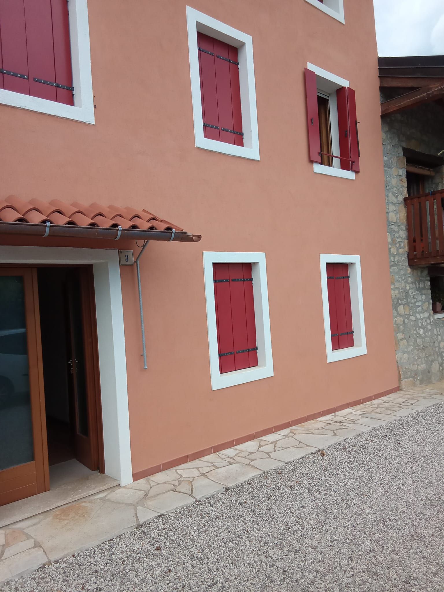 Casa color pesca con intonaco, persiane rosse, finiture bianche e cortile di ghiaia.