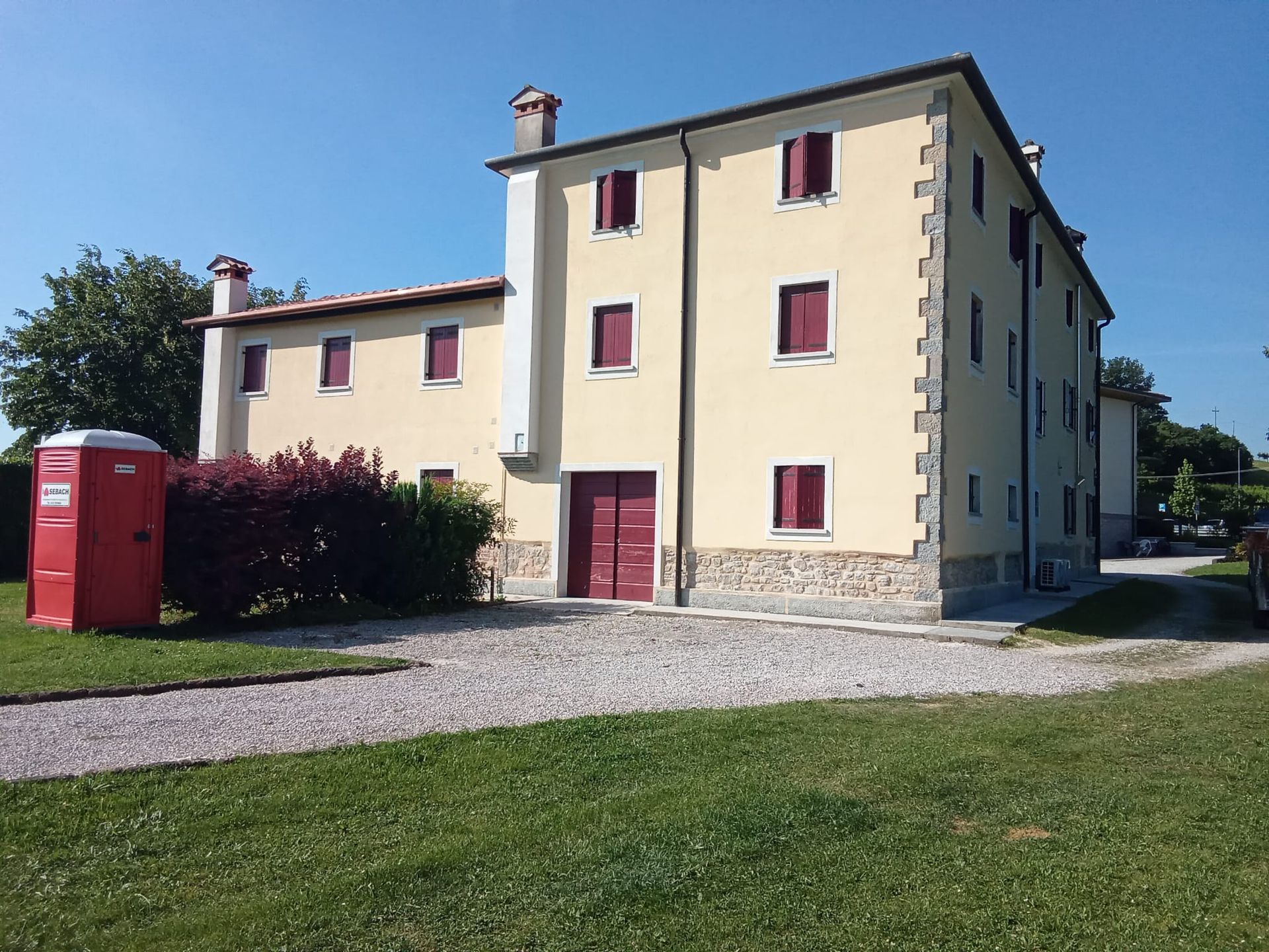 Palazzo di appartamenti giallo con persiane rosse, vialetto di ghiaia e cabina elettrica rossa in una giornata di sole.