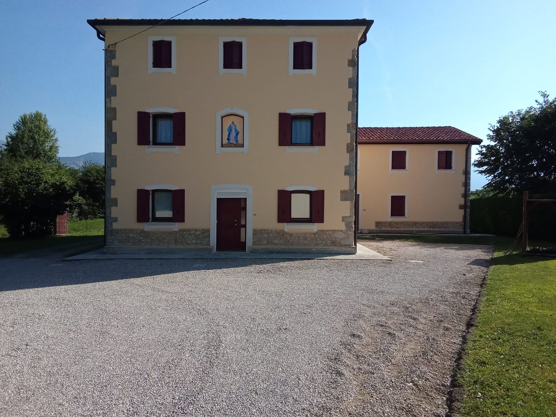 Ampio edificio beige con persiane rosse e un cortile di ghiaia sotto un cielo limpido.