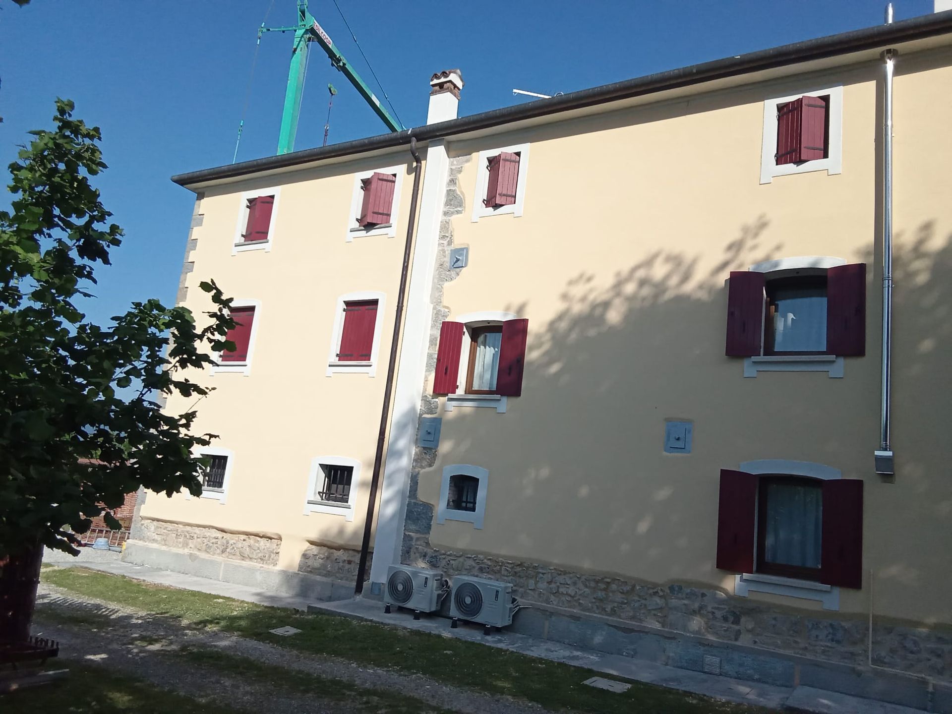 Palazzo di appartamenti color beige illuminato dal sole, con persiane rosse e un albero che proietta ombre sulla facciata.