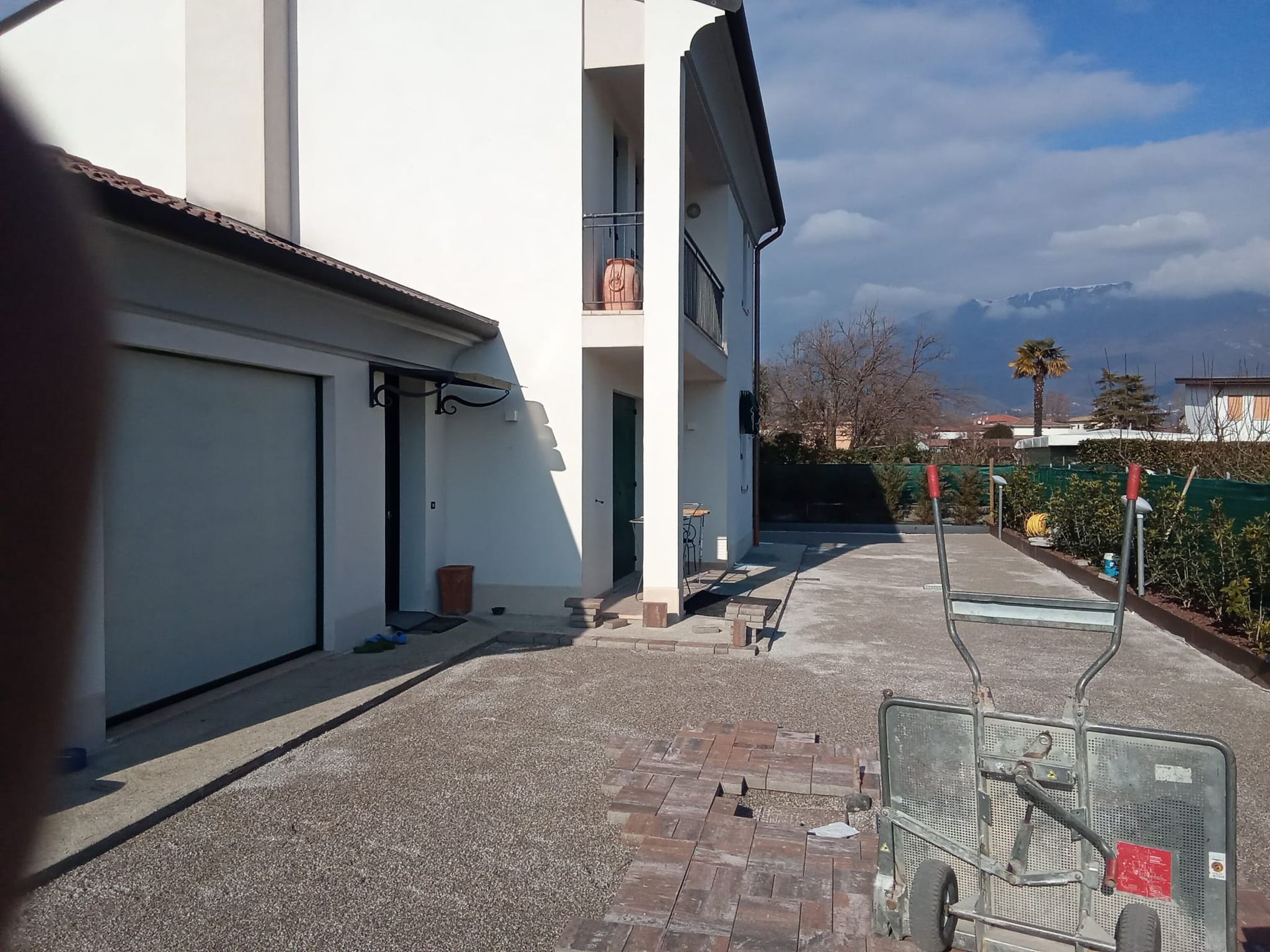 Vialetto residenziale soleggiato accanto a una casa bianca, con porta del garage, sentiero lastricato e cortile recintato sotto un cielo nuvoloso.