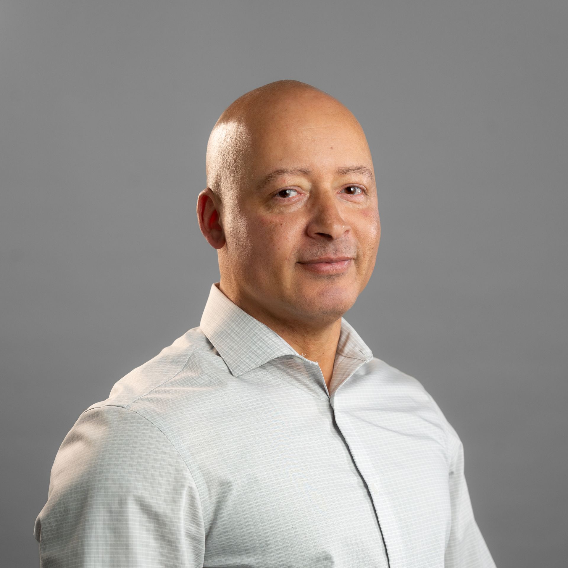 Bald man with tan skin in a light blue button-down shirt smiling at the camera. Grey background.