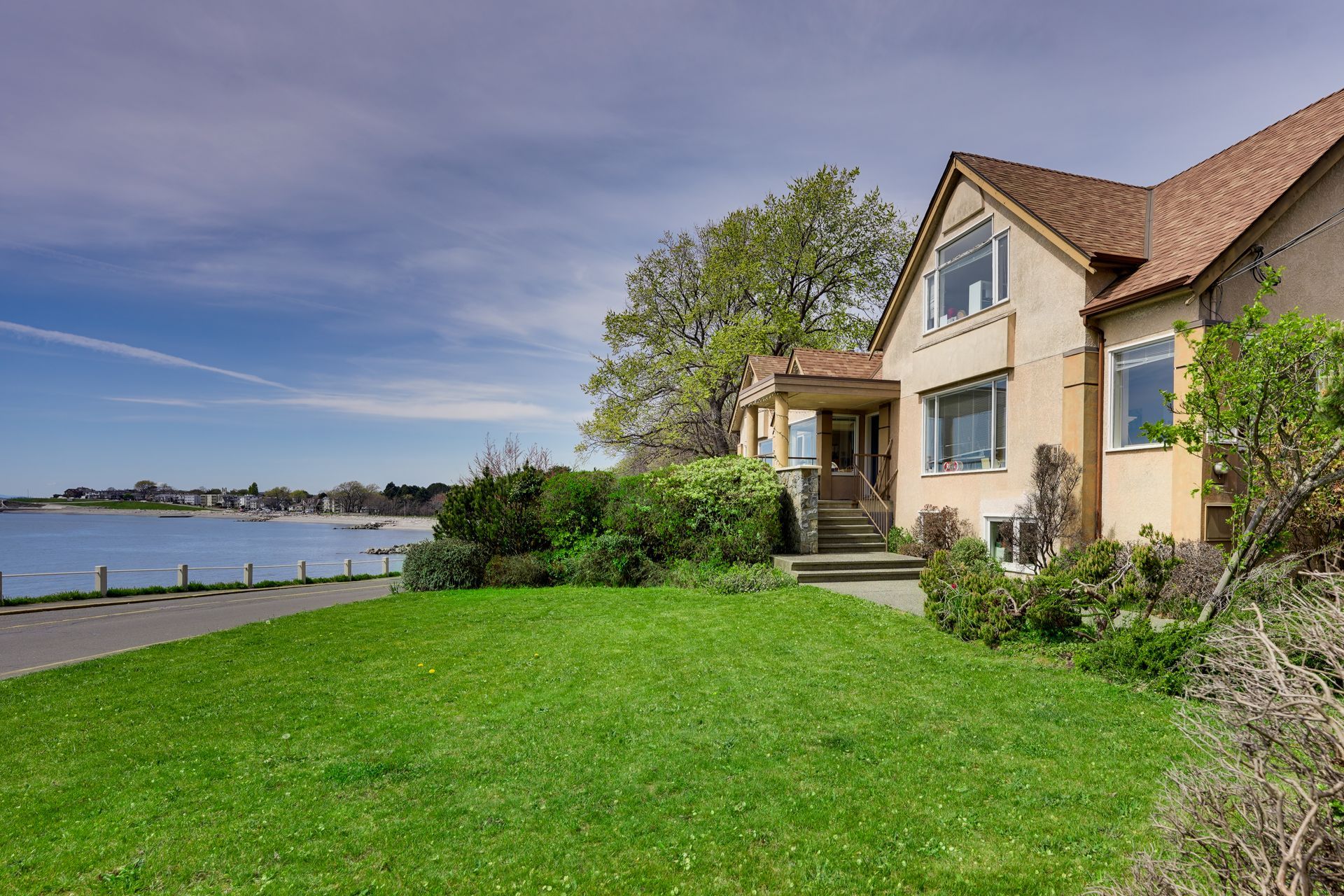 House with beige exterior, overlooking a blue water and a green lawn, on a sunny day.