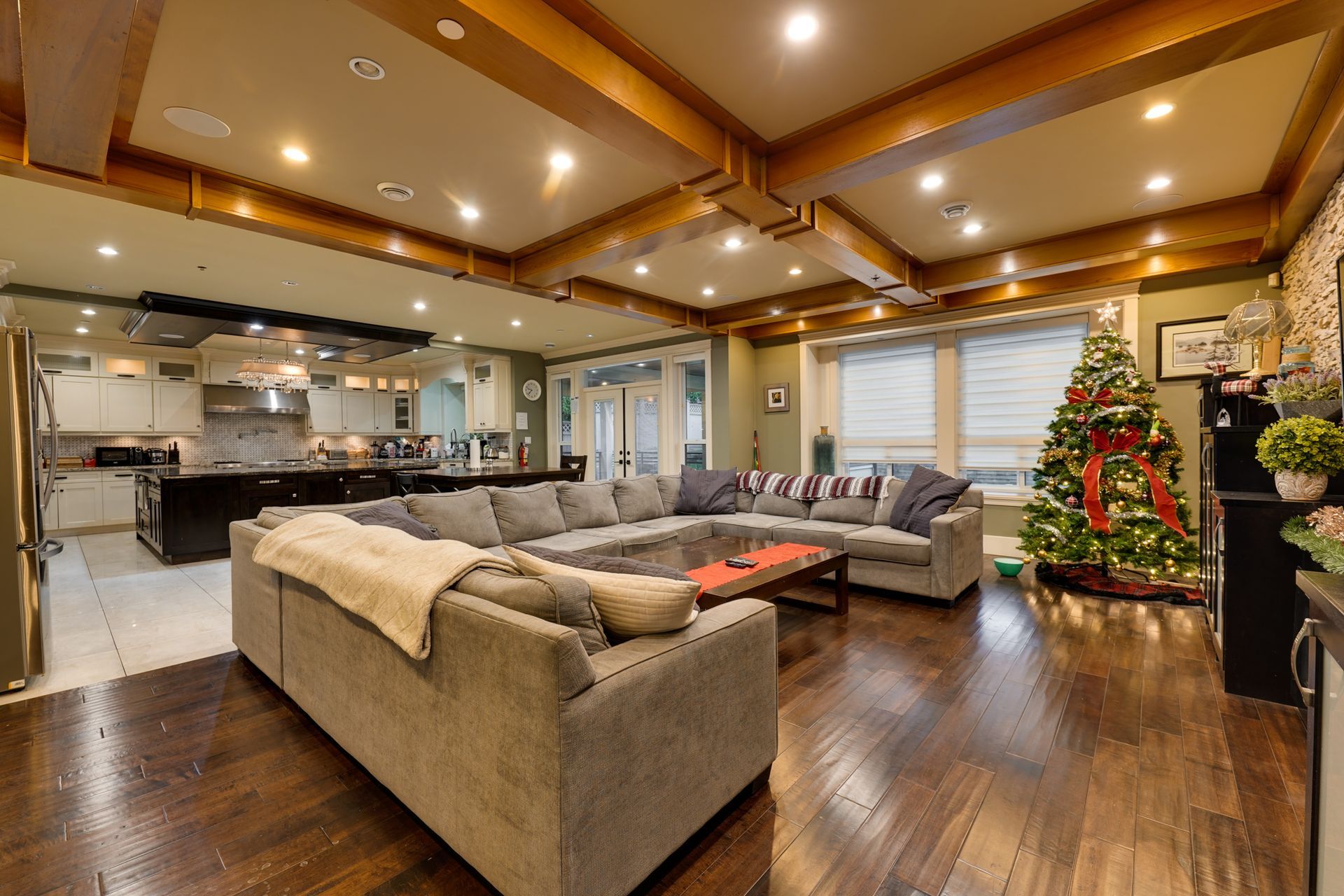 Living room with open kitchen; large gray sectional, Christmas tree, wooden beams, hardwood floor.