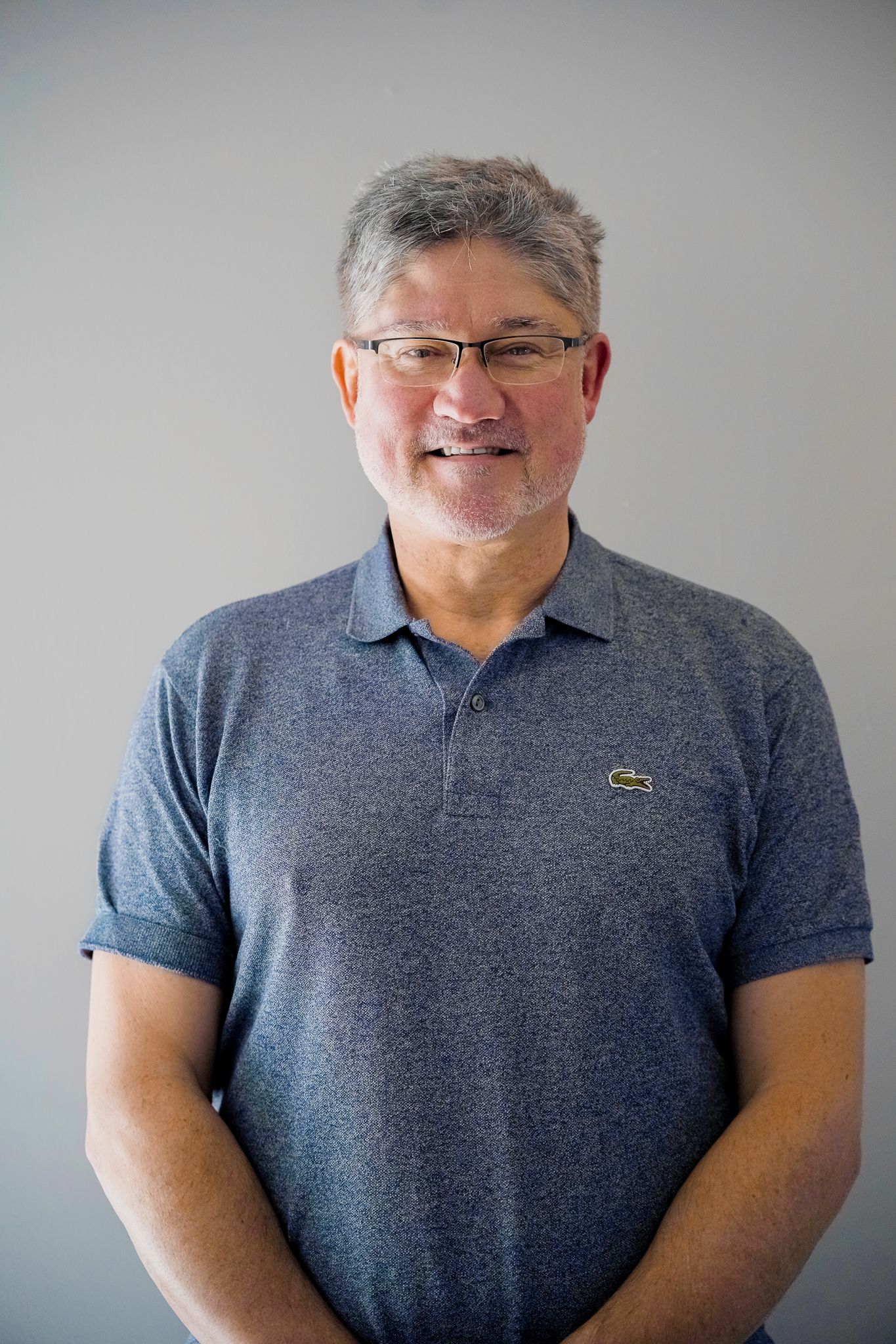 Man with glasses and salt-and-pepper hair wearing a blue polo shirt, smiling in front of a gray wall.