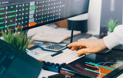 Hand using a keyboard at a desk with dual monitors showing stock charts and trading data