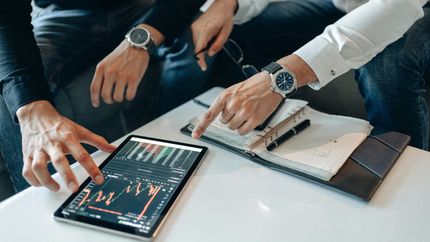 Two people reviewing stock charts on a tablet beside a notebook and phone on a desk