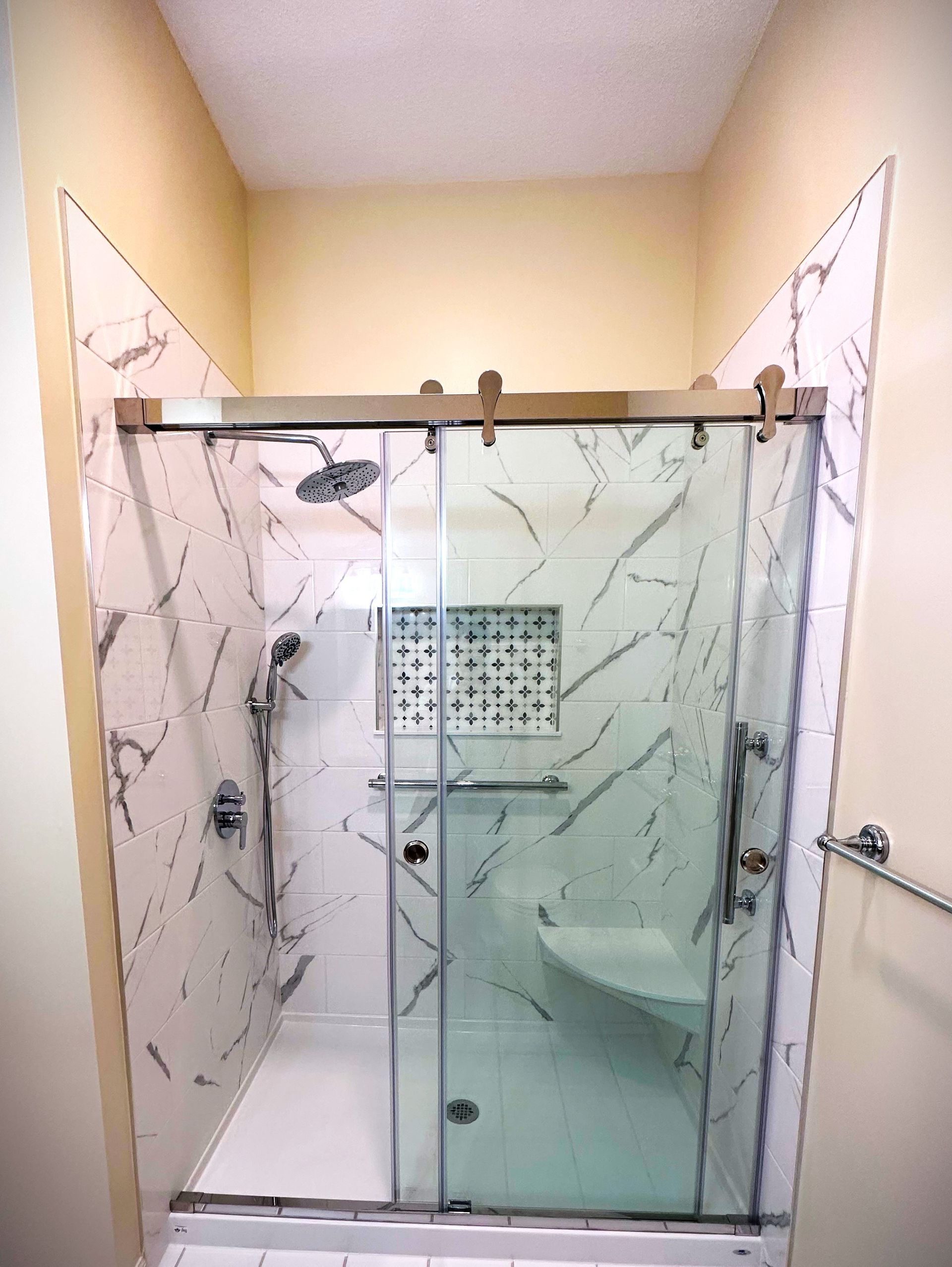 Modern shower stall with marble-like tile, glass doors, built-in seat, and a decorative tile niche.