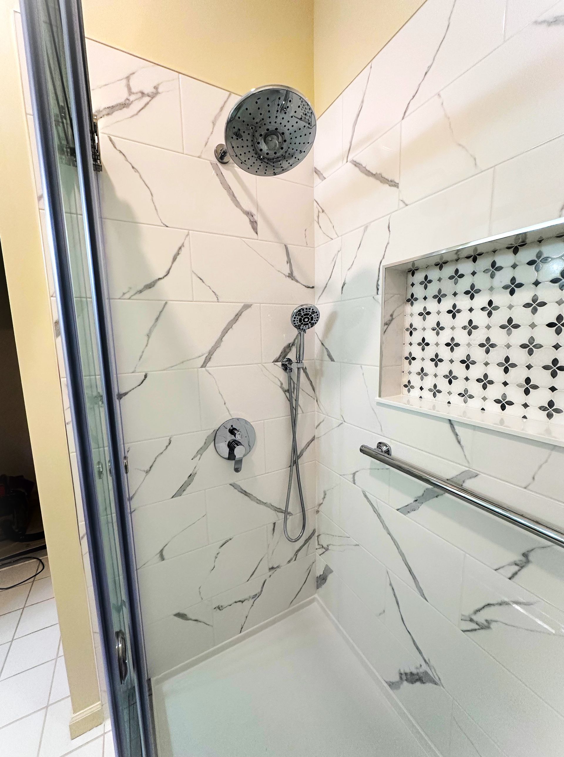 Shower with marble-look tile, rain shower head, handheld shower, recessed shelf, and a grab bar.