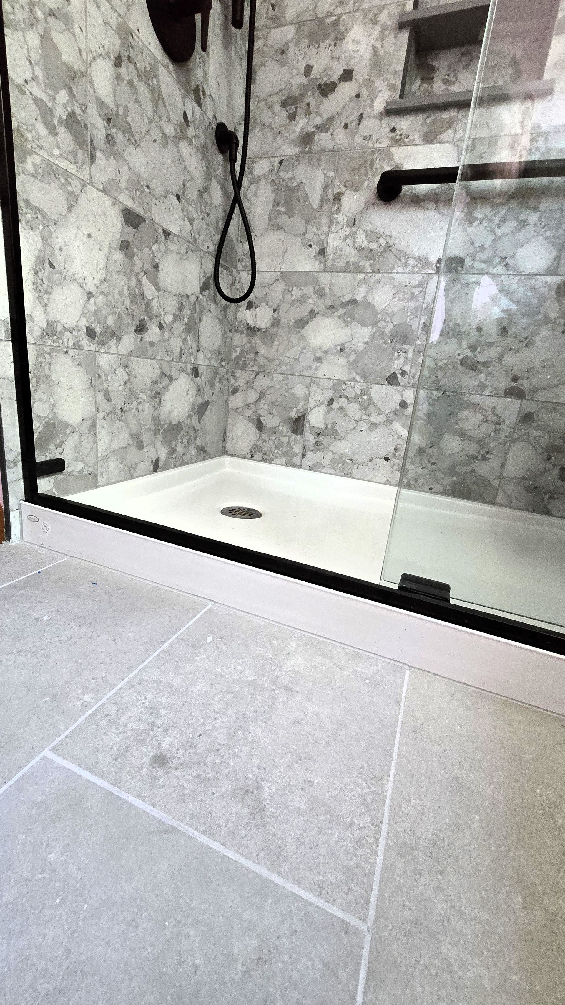 Shower with gray stone tile floor and walls. Black-framed glass door.