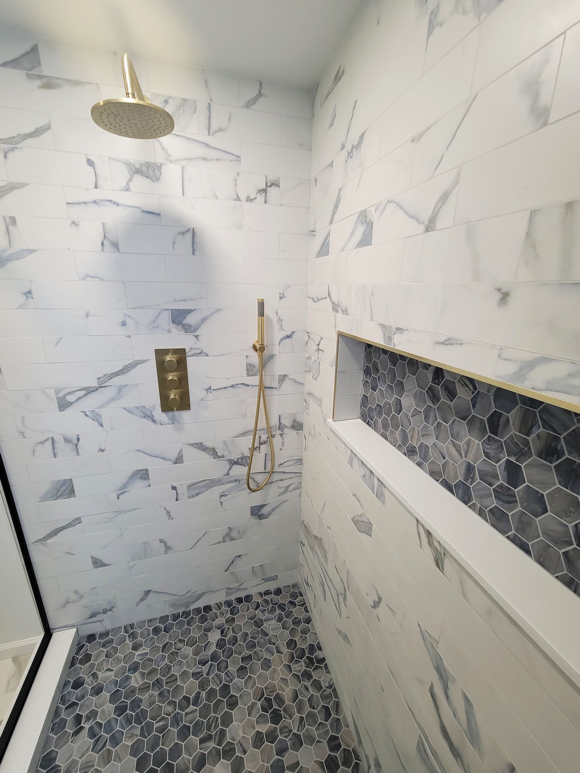Shower with gold fixtures and blue/white marbled tile. Pebble floor and a built-in shelf with dark mosaic tile.