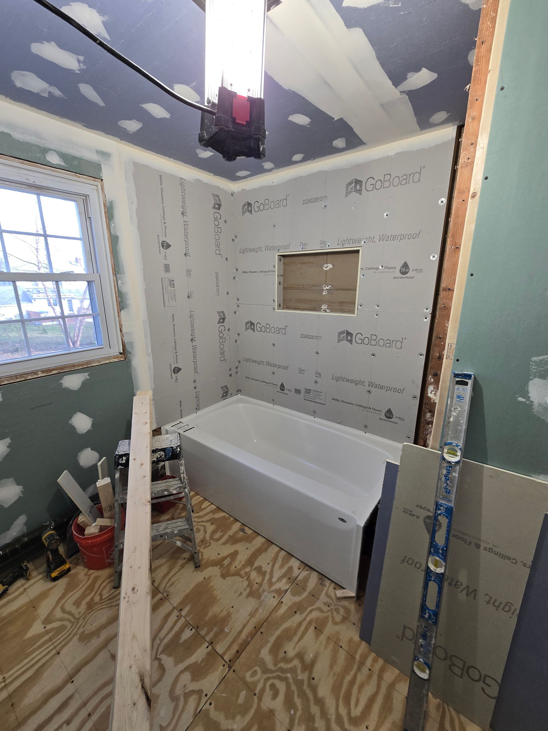 Bathroom renovation: walls with cement board, tub, window, plywood floor, ladder, and tools.
