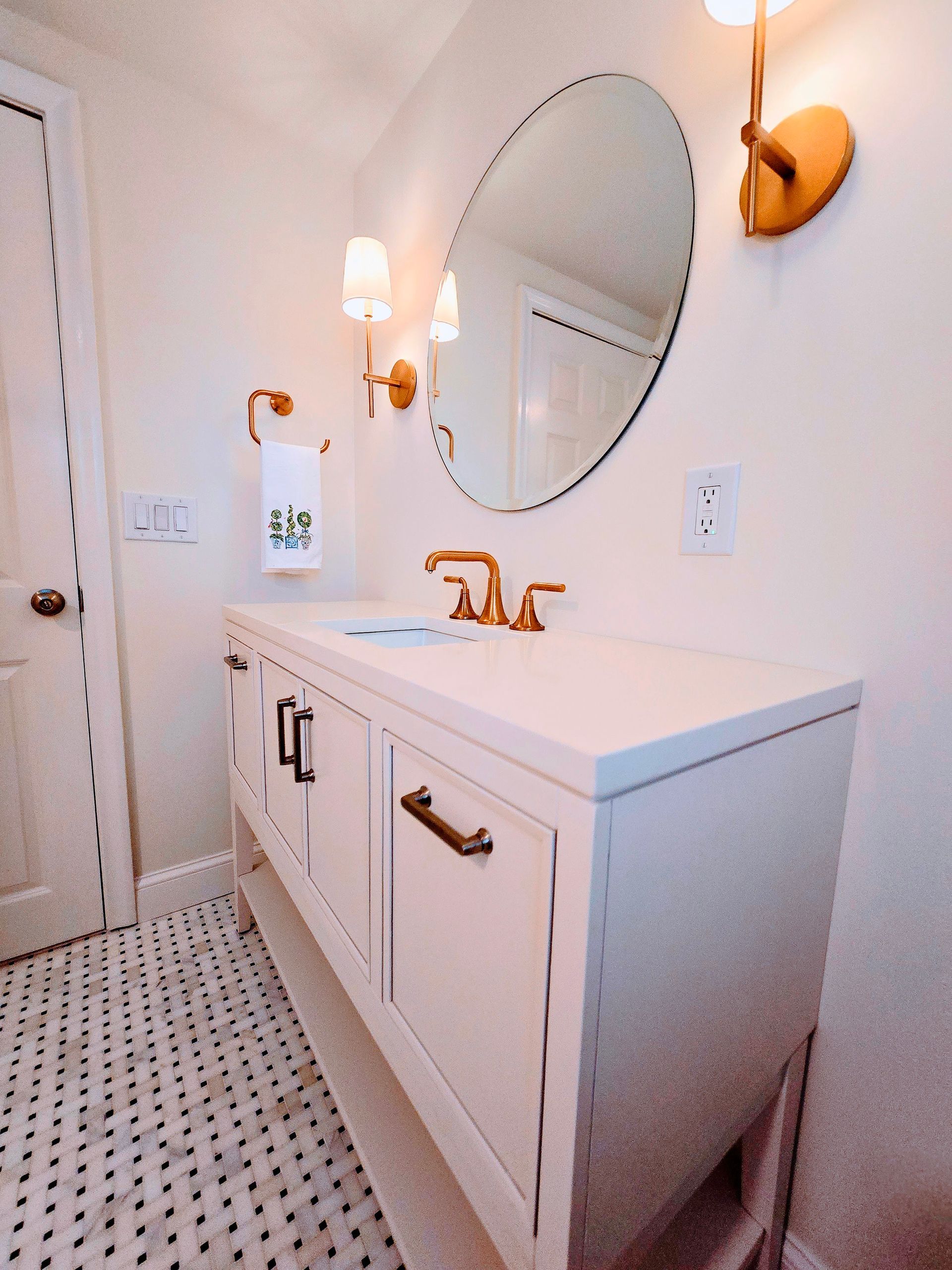 White bathroom with gold fixtures, oval mirror, white vanity, and patterned floor.