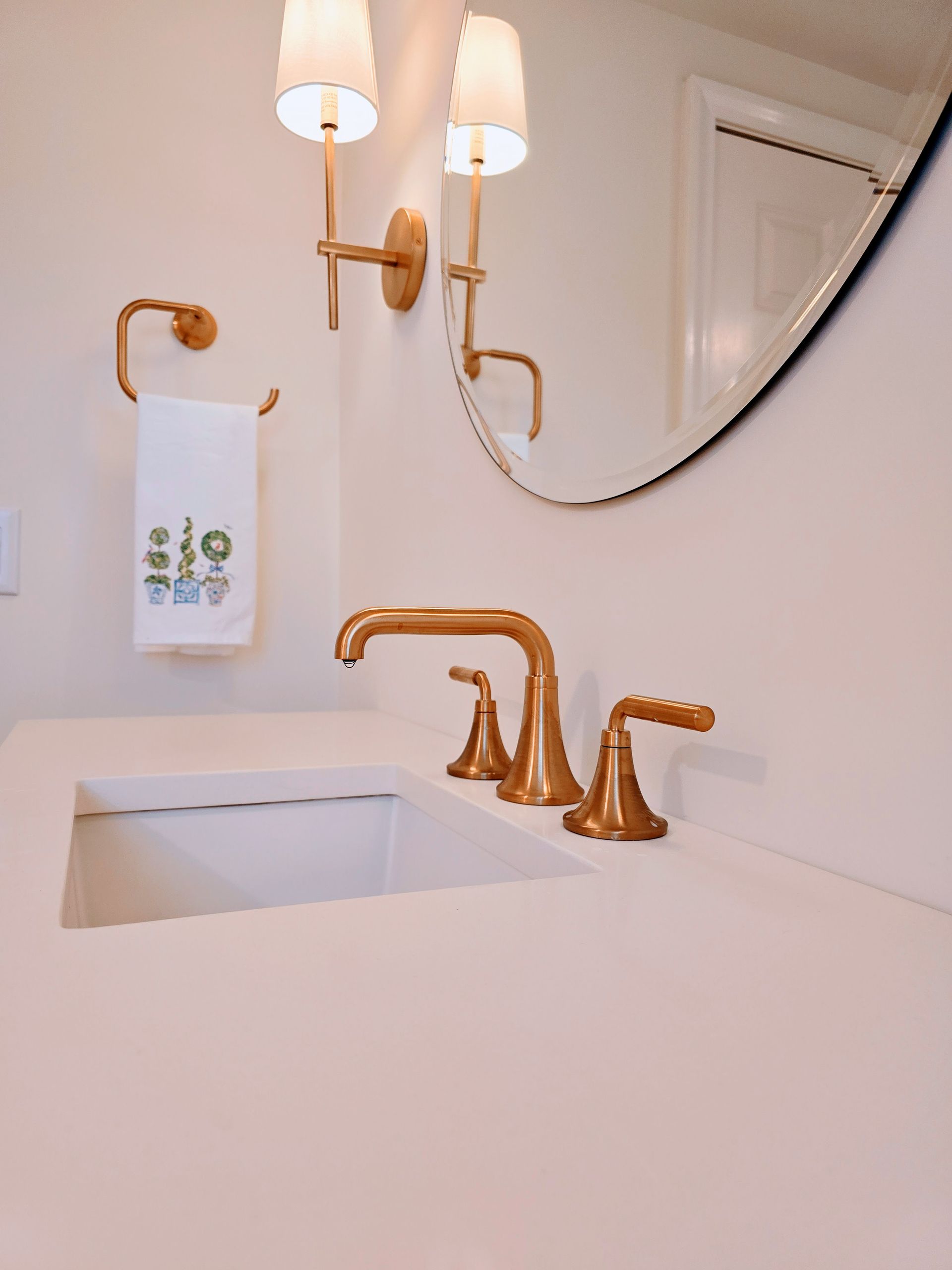 Gold fixtures adorn a white bathroom sink and countertop. Towel with plants hangs on a wall, with a round mirror and sconces above.