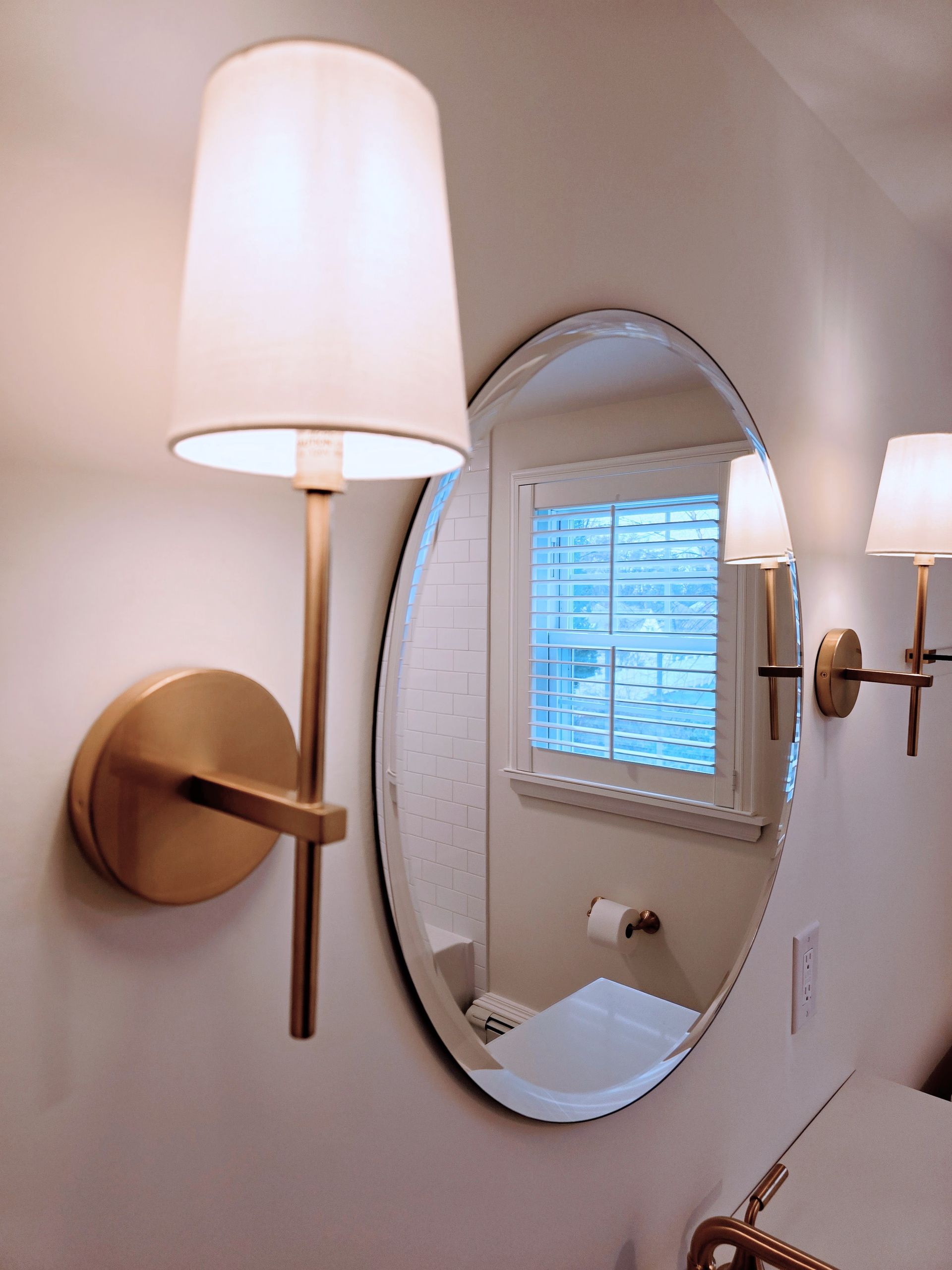 Two gold sconces with white shades flank an oval mirror. Window and sink visible.