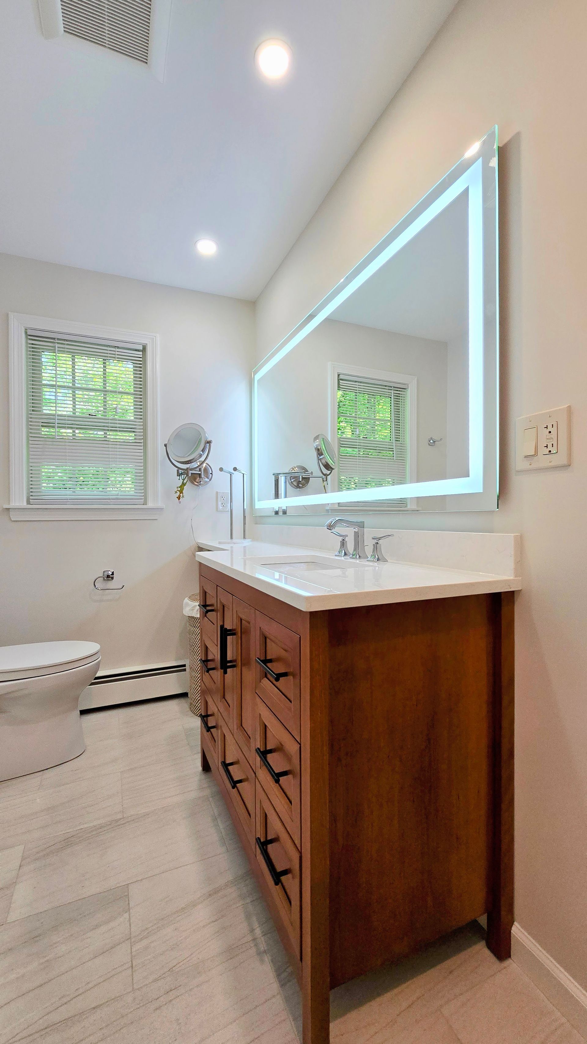 Bathroom with wooden vanity, illuminated mirror, and toilet. Light-colored walls and floor, window with blinds.