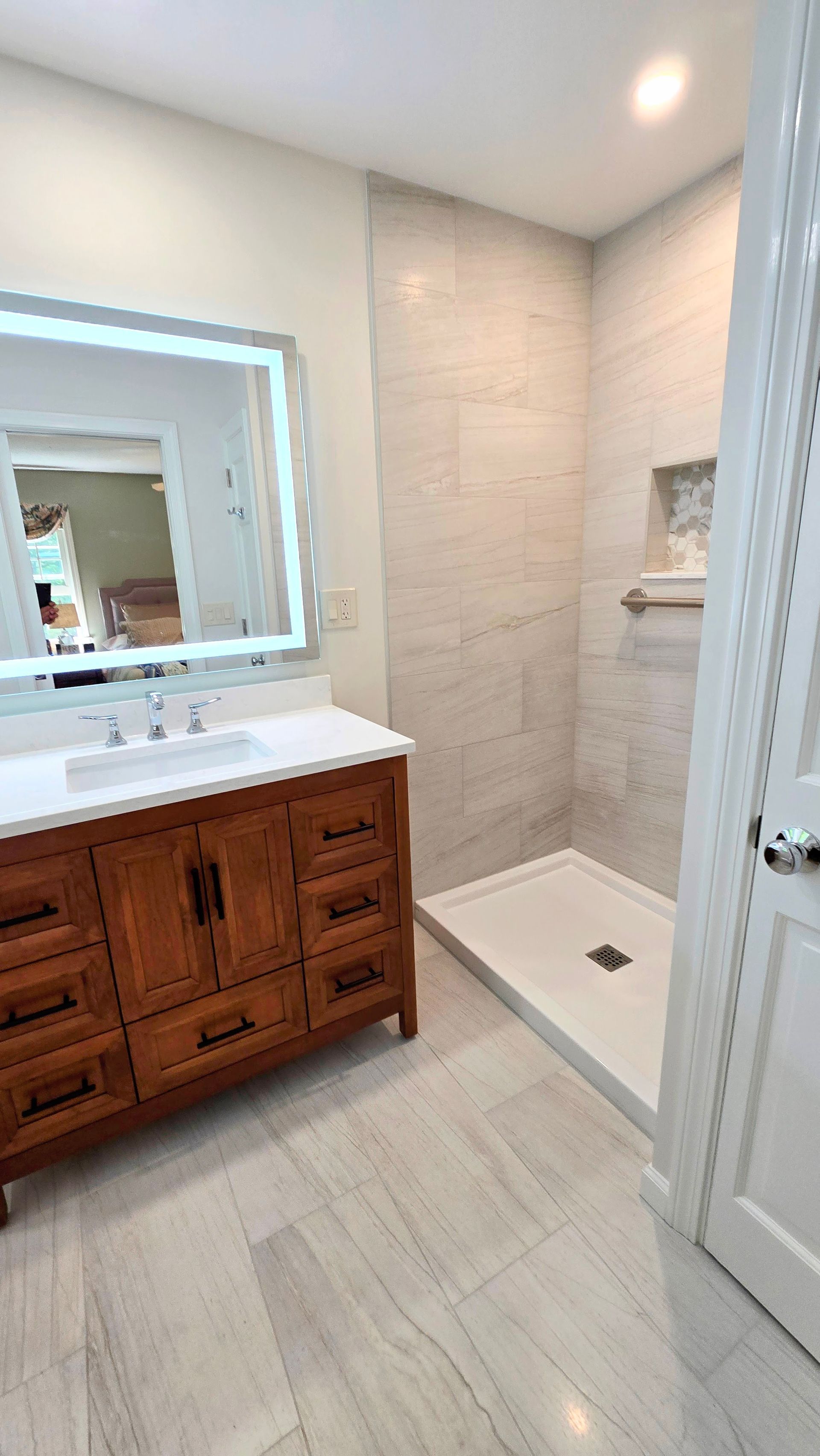 Bathroom with wooden vanity, large lit mirror, tiled shower, and light-colored tile floor and walls.