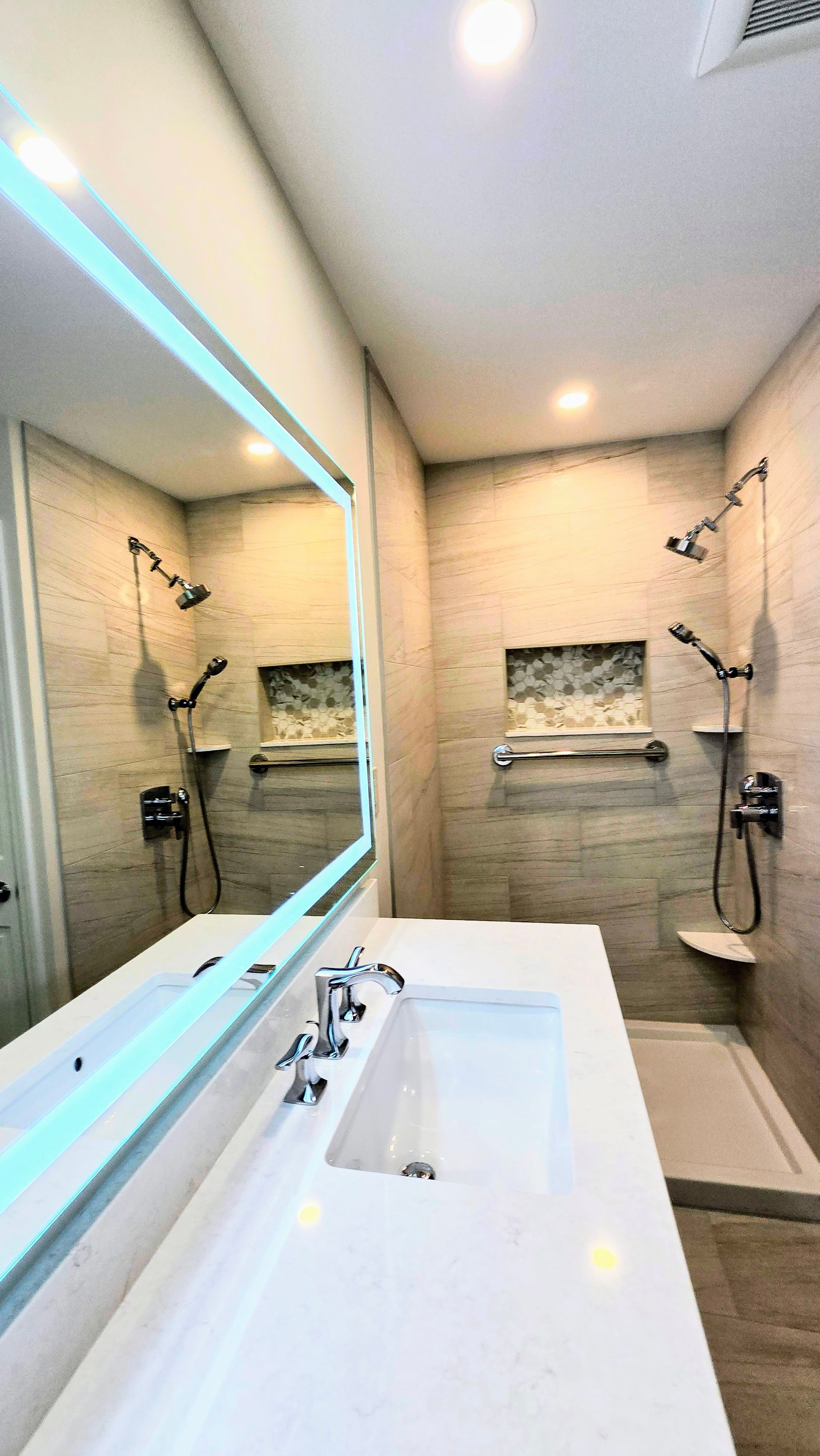 Bathroom with a white countertop, large mirror, and two shower heads. Light beige tile walls.