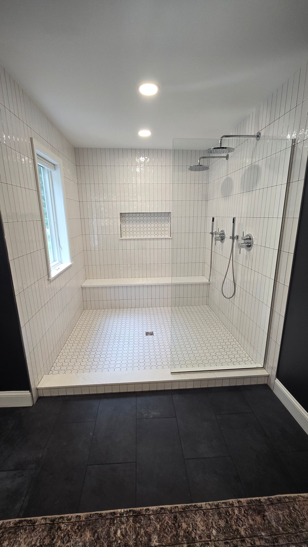 A modern bathroom with a large shower, white tiled walls, glass door, and dark floor.