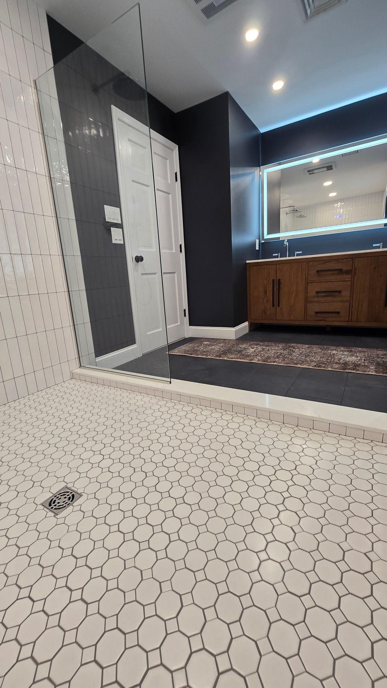 Modern bathroom with white pebble-tile floor, glass shower, dark walls, wooden vanity, and illuminated mirror.