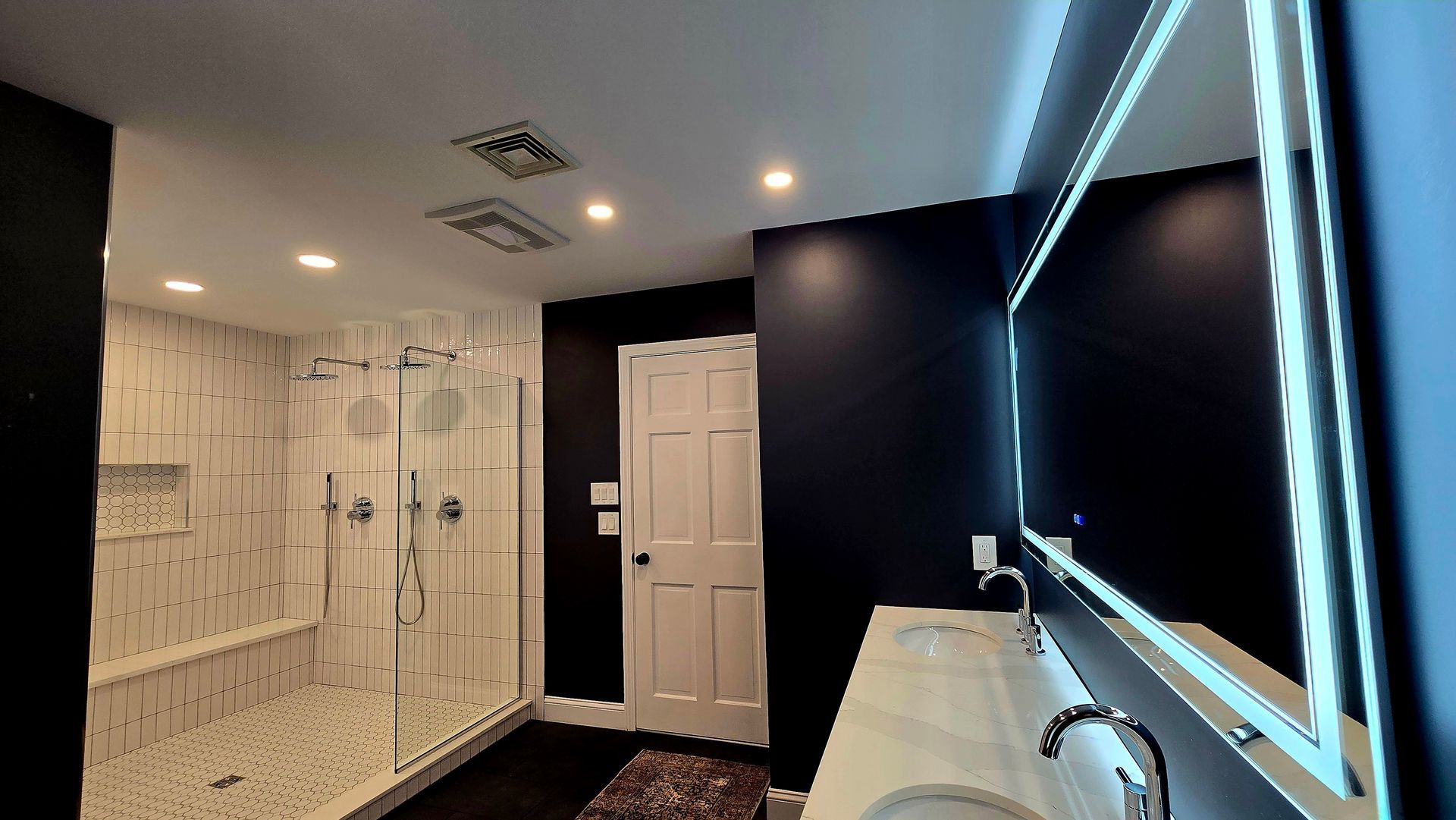Modern bathroom with glass shower, dark blue walls, white vanity, and large illuminated mirror.