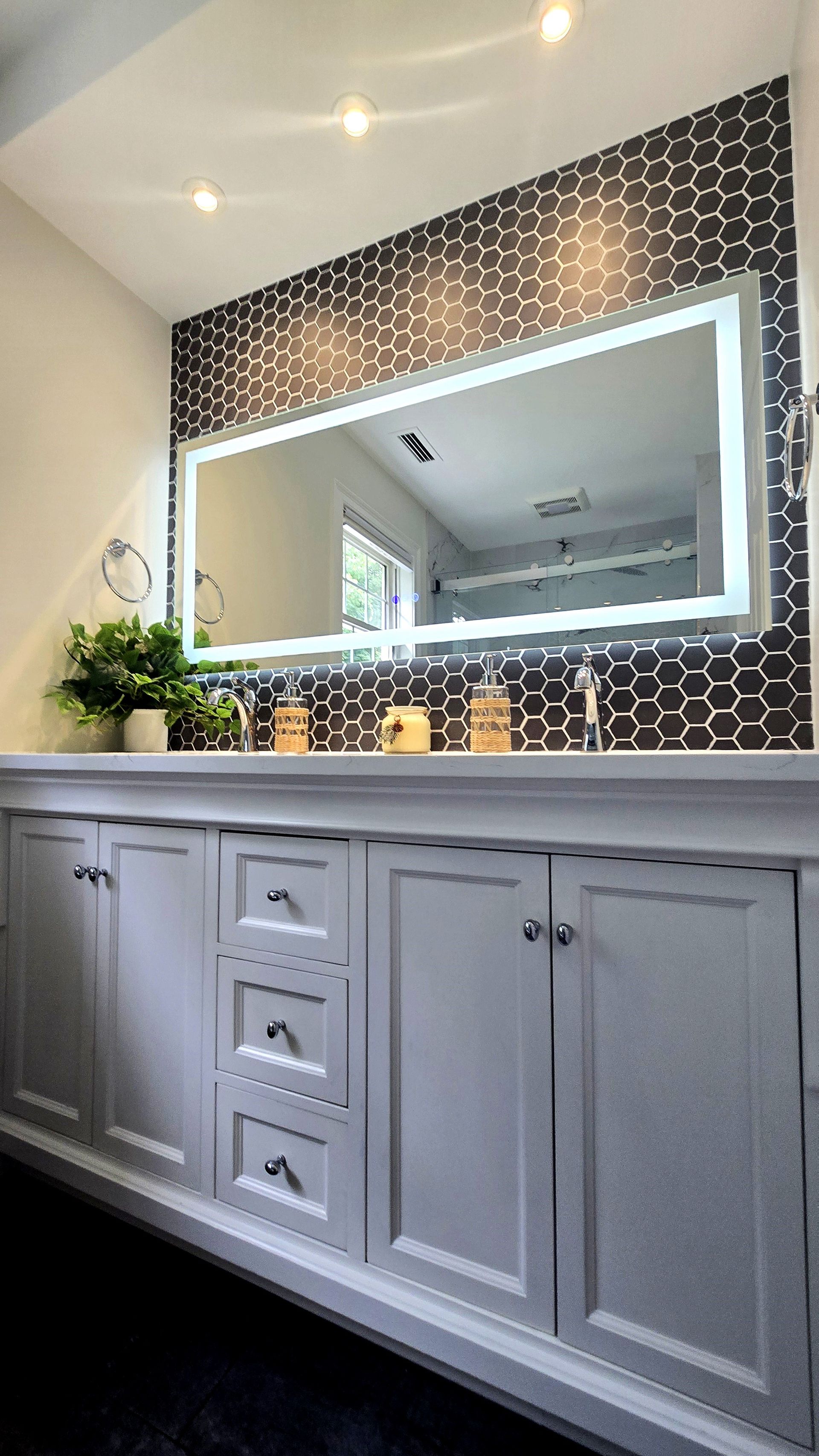 Bathroom vanity with lit mirror in Farmington, CT with dark tiled accent wall, and white cabinetry.