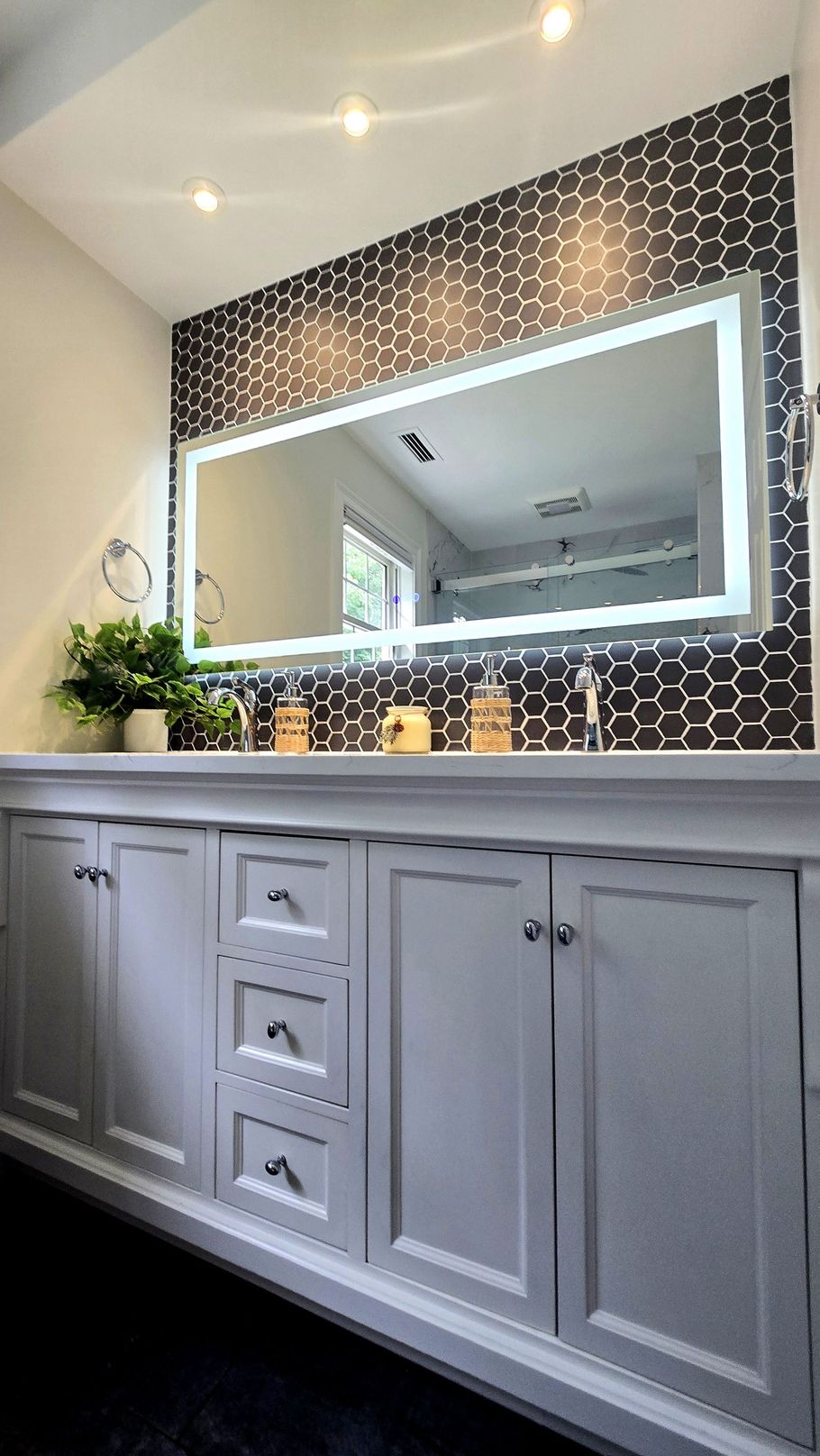 Bathroom vanity with lit mirror in Farmington, CT with dark tiled accent wall, and white cabinetry.