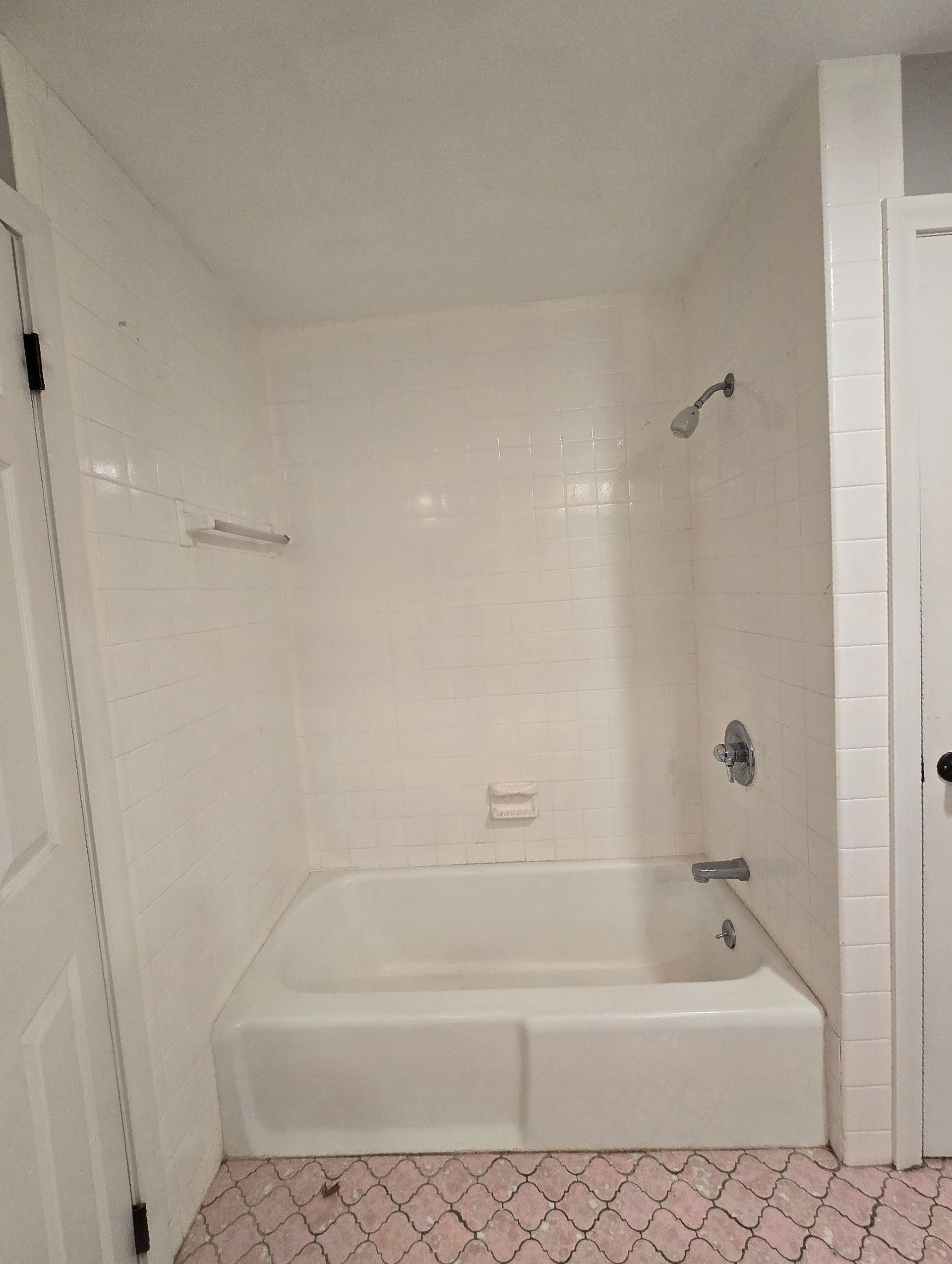 White-tiled bathroom with bathtub, showerhead, and pink patterned floor.