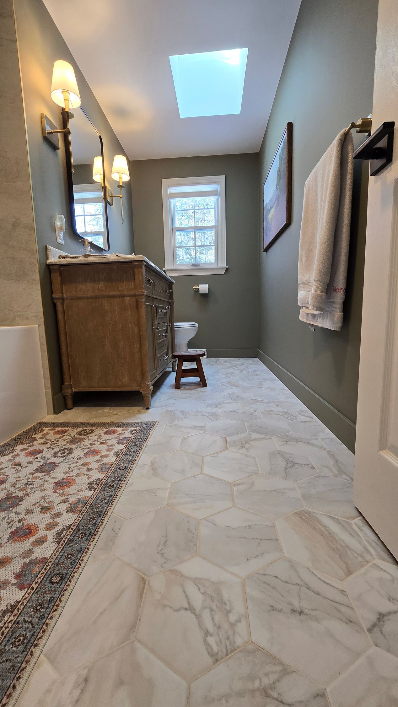 Bathroom with marble tile floor, olive green walls, wooden vanity, and skylight.