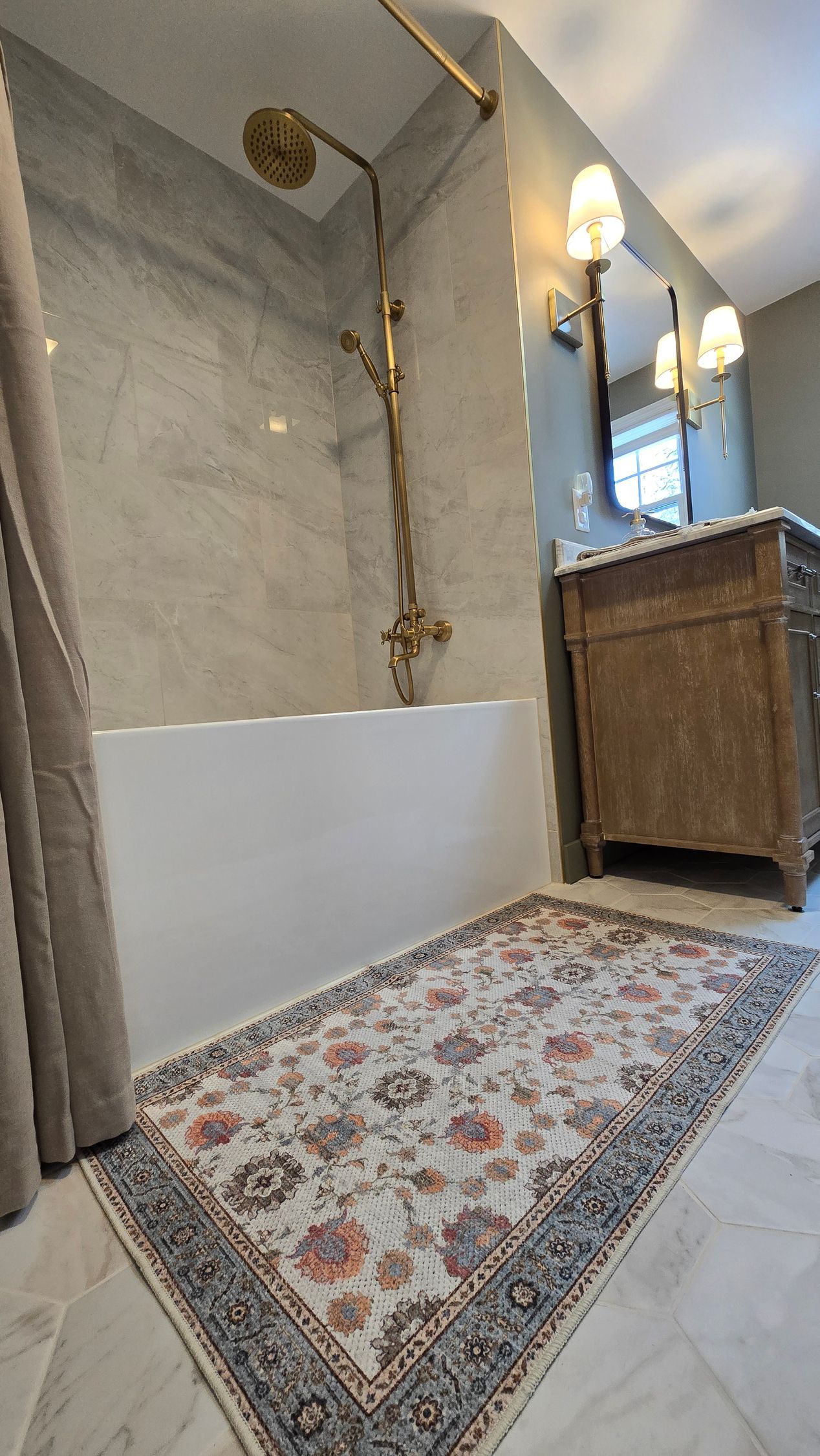 Bathroom with gold shower fixture, white tub, patterned rug, and wooden vanity.