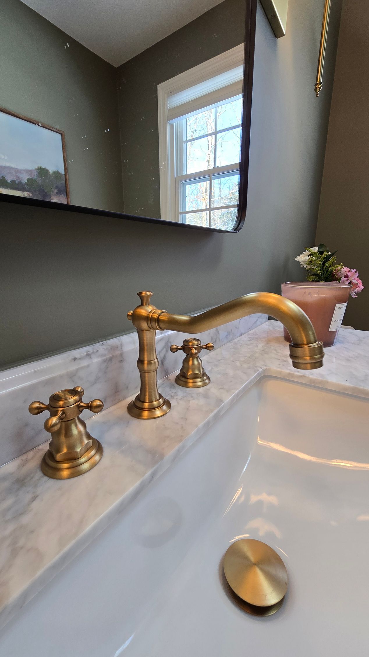Gold faucet on a white marble countertop in a bathroom, with a mirror and a window in the background.