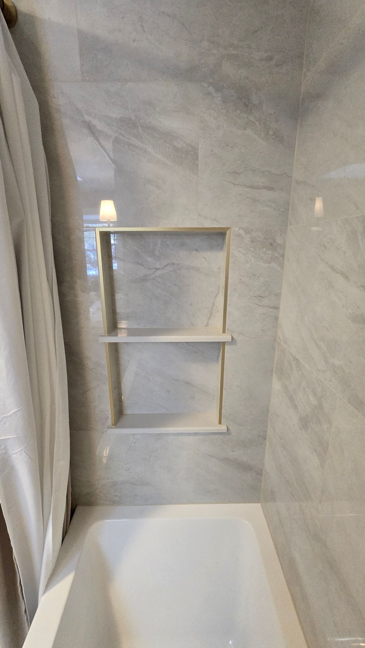 Bathtub with built-in shower shelves in a marble-look tiled alcove.