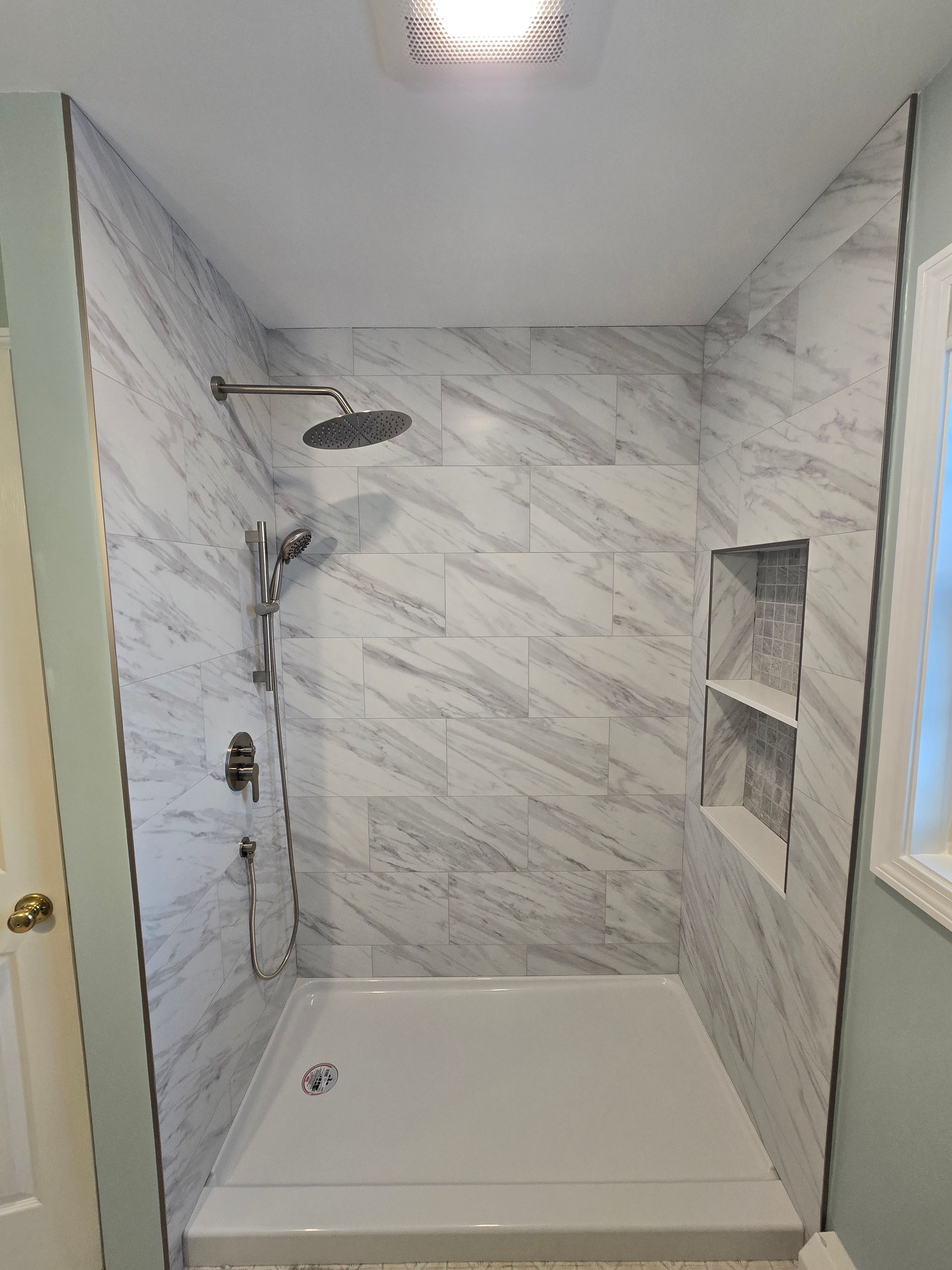 Tiled shower stall with rain showerhead and hand shower. Marble-like wall tiles, white shower base.