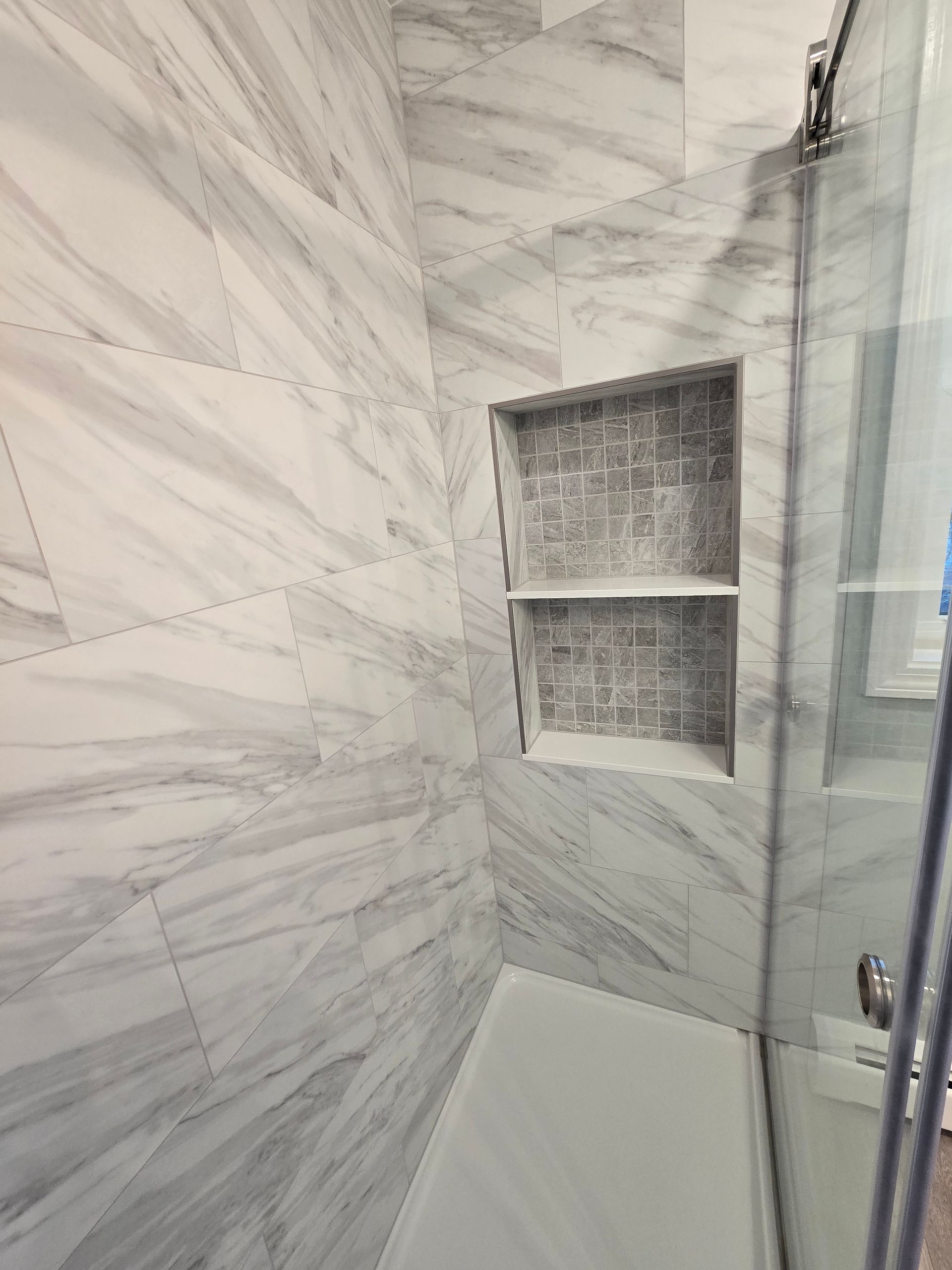 Shower with marble-look tile, a recessed shelf, and a glass door. White and gray tones dominate the interior.