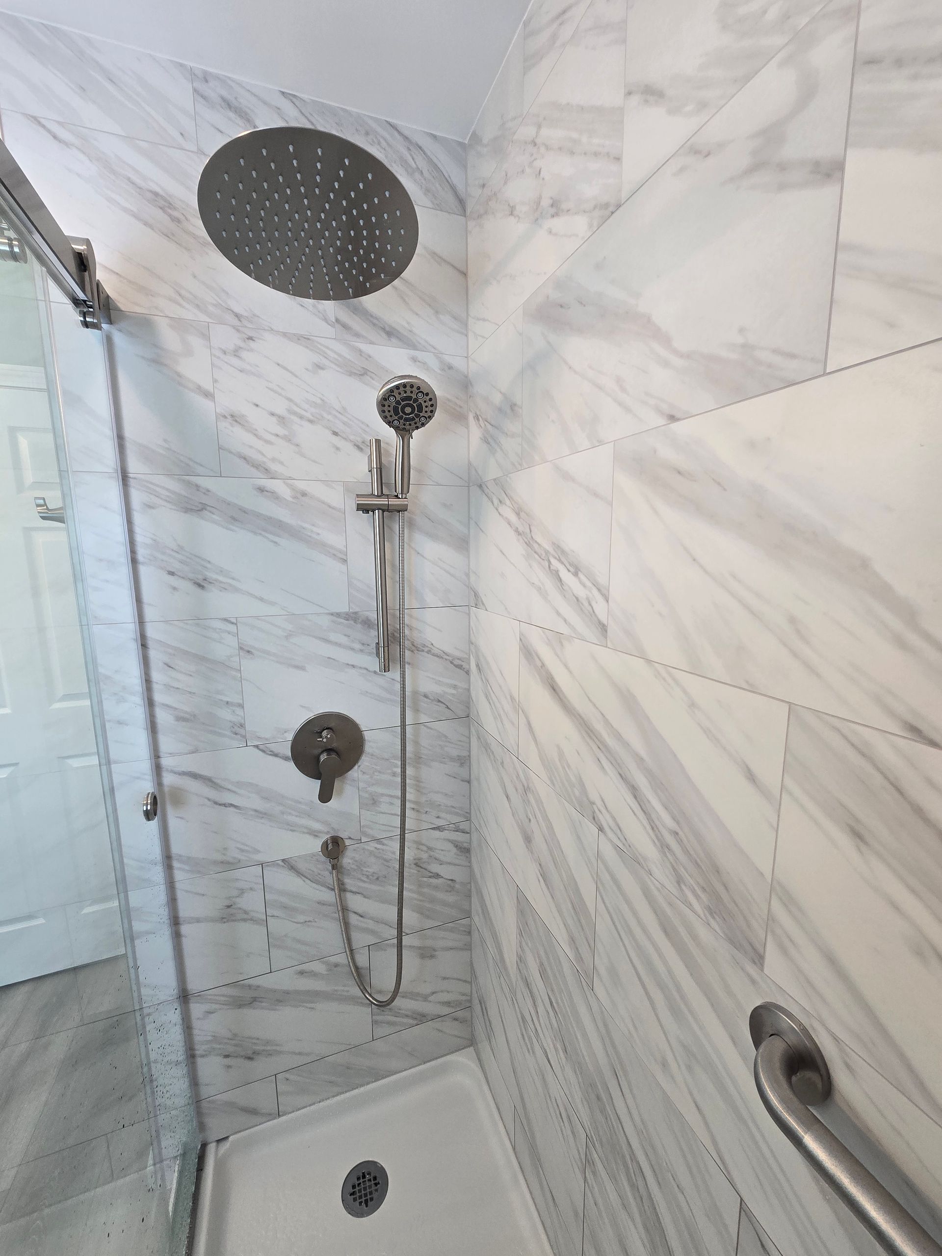 Shower with marble-look tile, rain shower head, hand shower, and grab bar for accessibility.