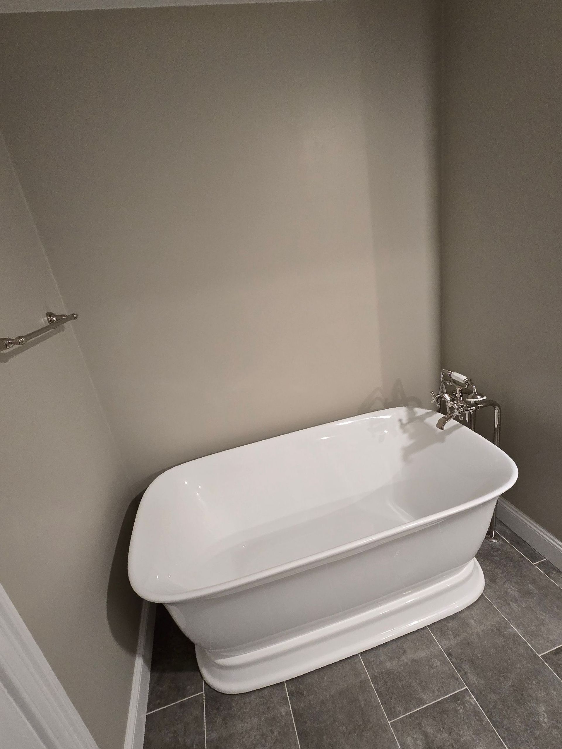 White freestanding bathtub in corner of bathroom with gray tile floor and walls.