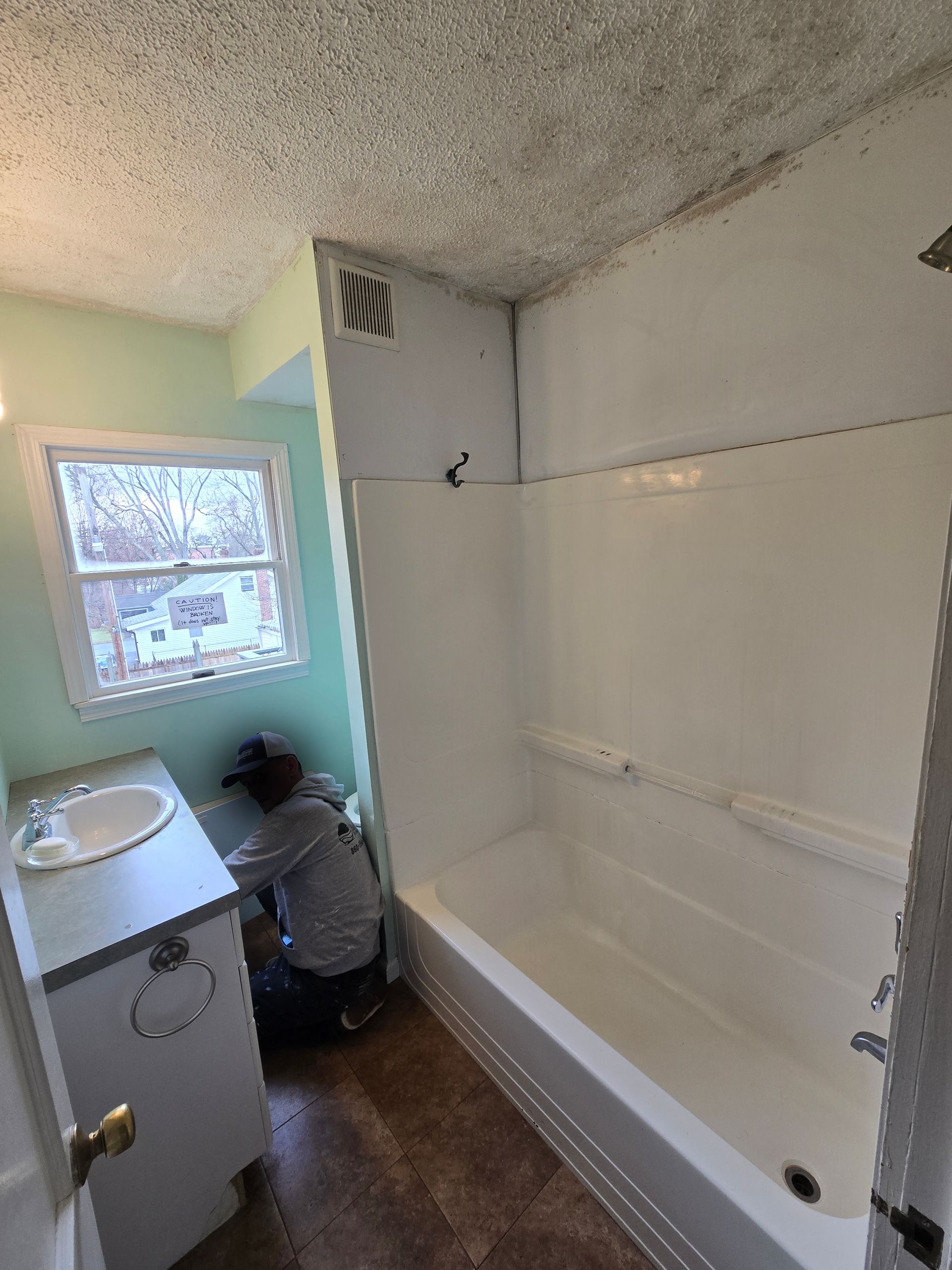 A person works on plumbing in a bathroom with a white shower/tub, vanity, and window.