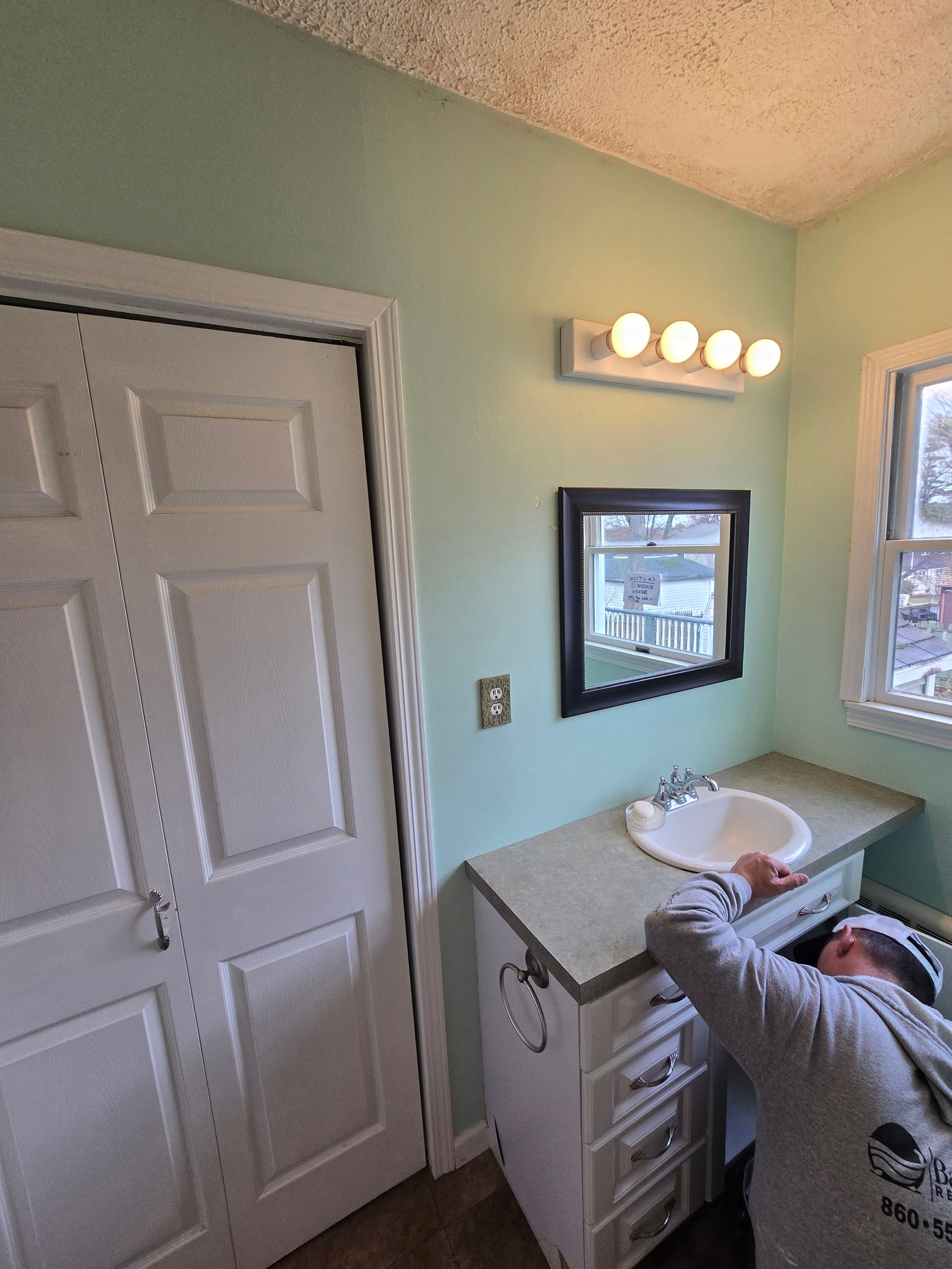 A person working on bathroom vanity; mint green walls, white cabinets, four-bulb light fixture above a black-framed mirror.