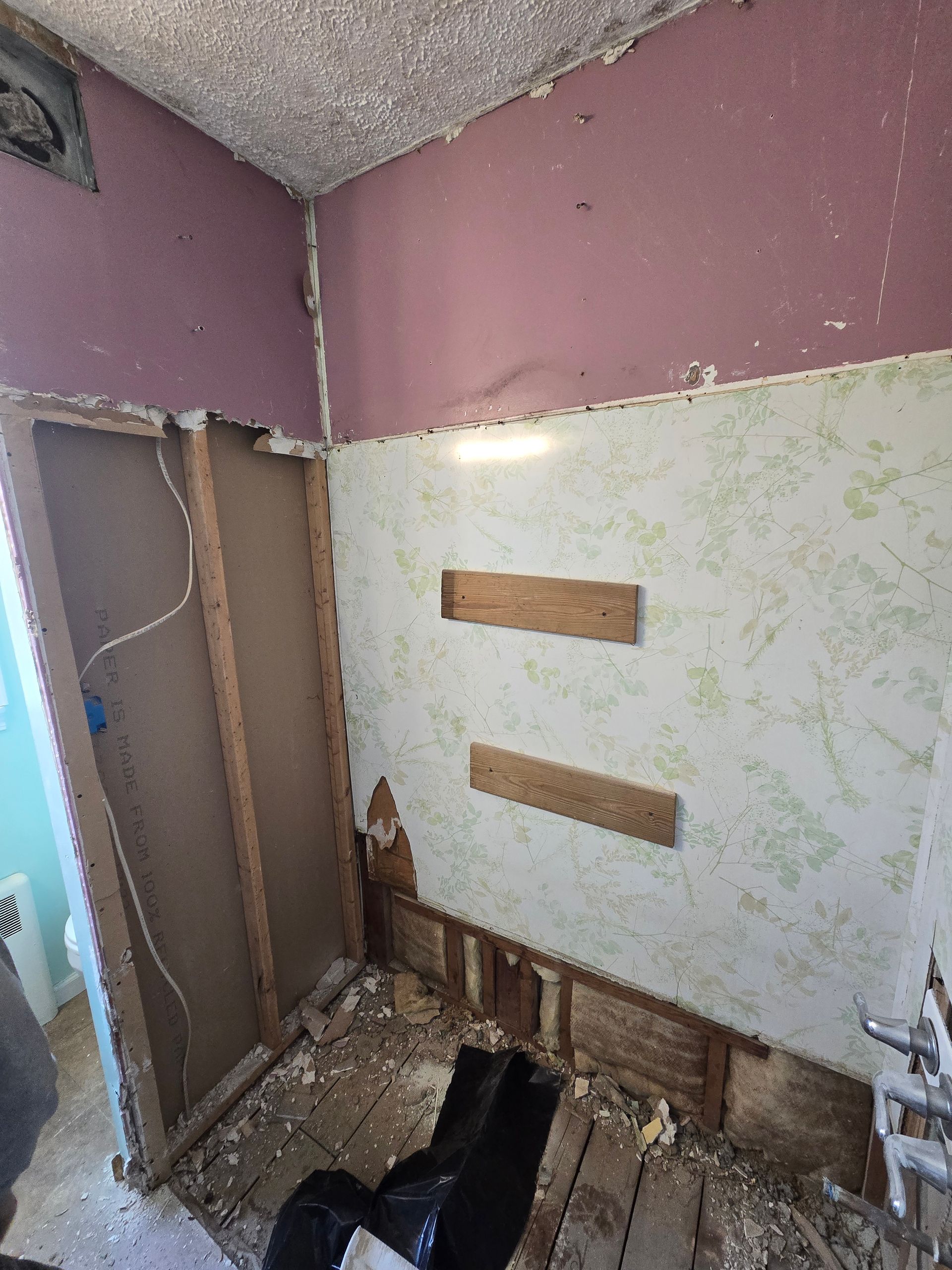 Bathroom under renovation: exposed walls, pink and patterned wallpaper, wood framing, debris on floor.