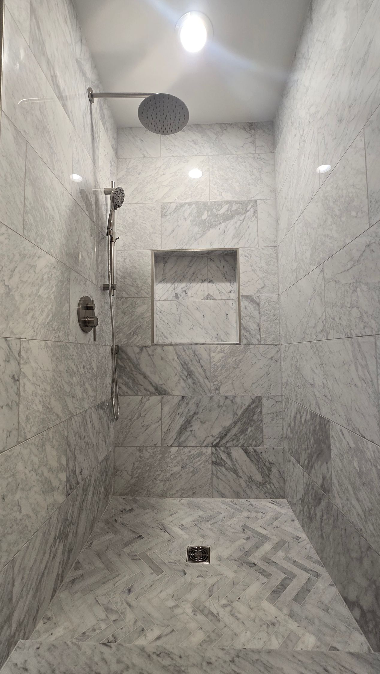Marble-tiled shower with rainfall showerhead, handheld sprayer, recessed shelf, and herringbone floor pattern.