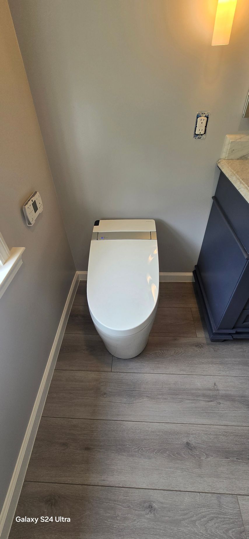 A modern toilet in a small bathroom with gray walls and wood-look flooring.