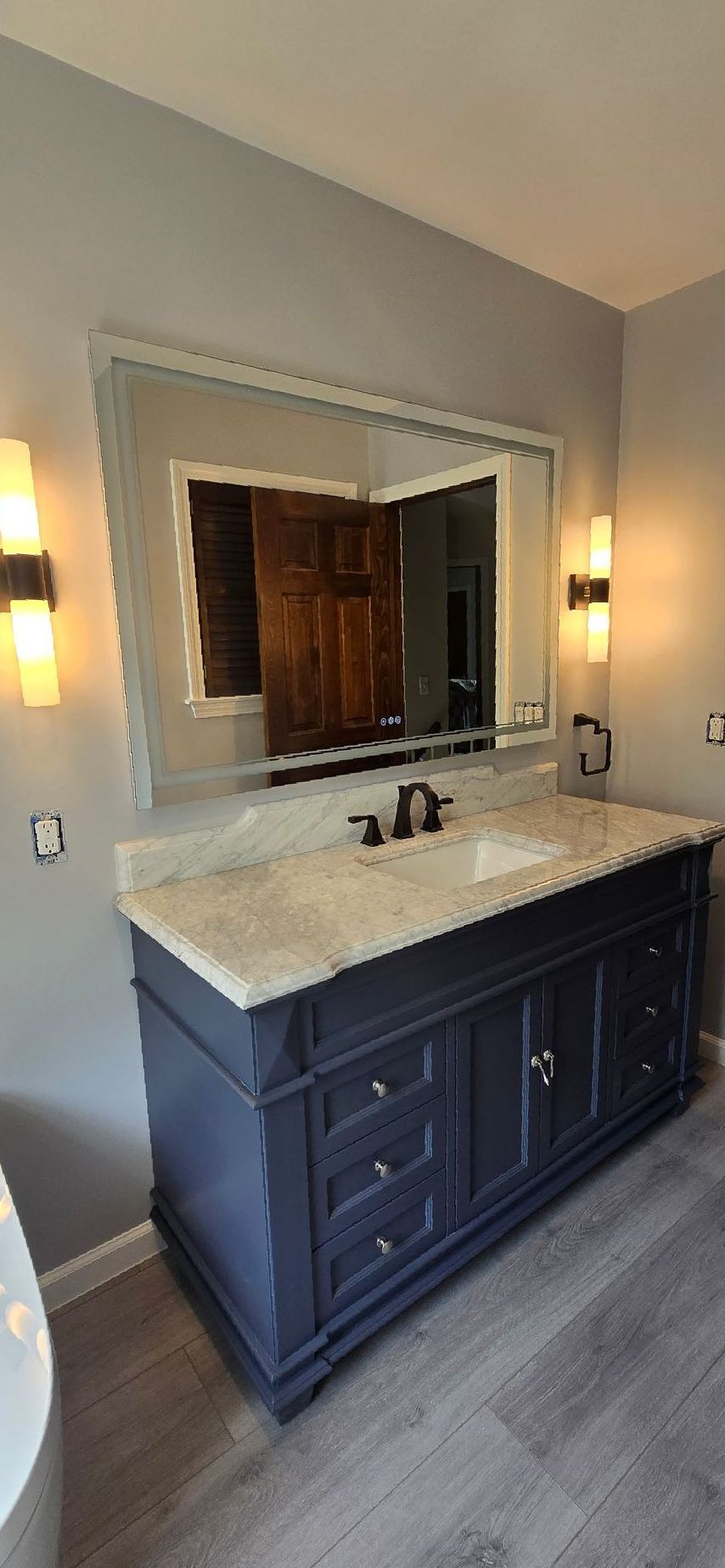 Bathroom vanity with a dark blue cabinet, light-colored countertop, large mirror, and sconces on a grey wall.
