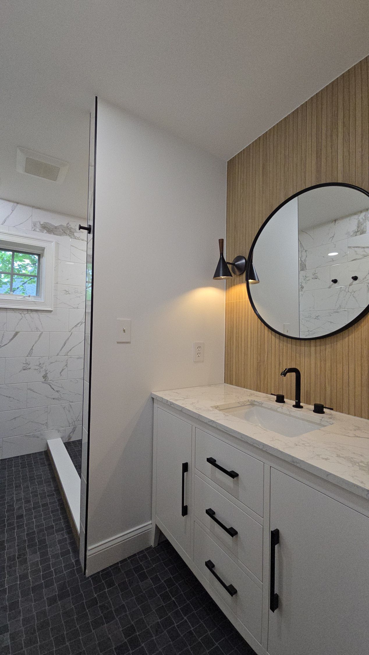 Bathroom with white vanity, black fixtures, round mirror, and wooden wall accent.
