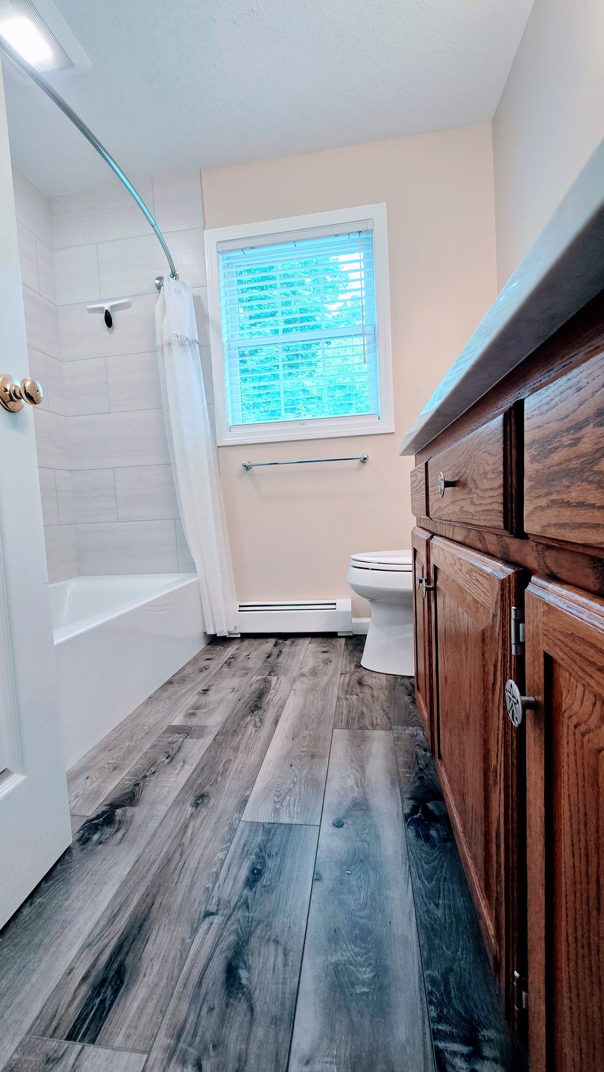 Bathroom with wood-look floor, shower, vanity, toilet, and window with blinds; neutral colors.