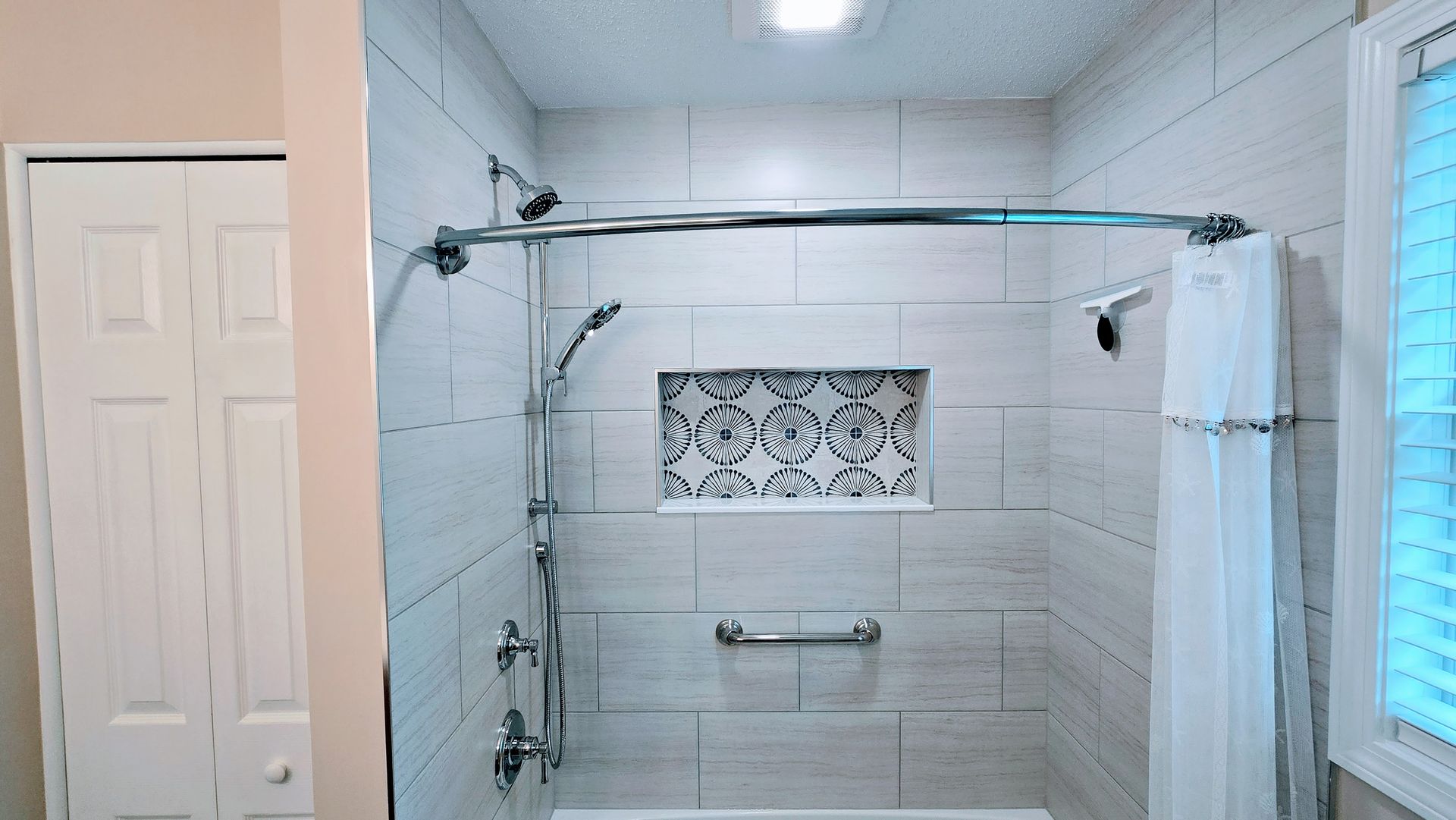White tiled shower with a built-in shelf and chrome fixtures. White shower curtain, grab bar.